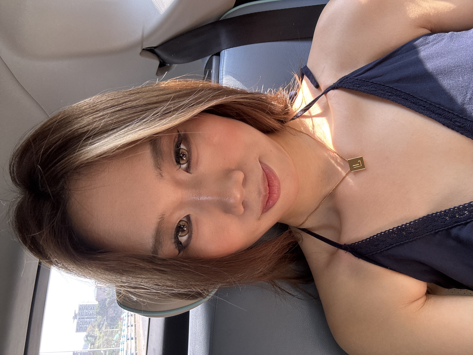 A young woman poses for a selfie in the backseat of a car. She has shoulder-length brown hair with blonde highlights and is wearing a dark blue tank top with lace trim and a gold necklace with a rectangular pendant. Her makeup includes winged eyeliner and blush. Outside the car window, a high-rise building and green foliage can be seen, suggesting an urban environment during daylight hours. The location can be narrowed down to Nan'an District, China. The presence of a seatbelt suggests the car is likely in motion or recently stopped.