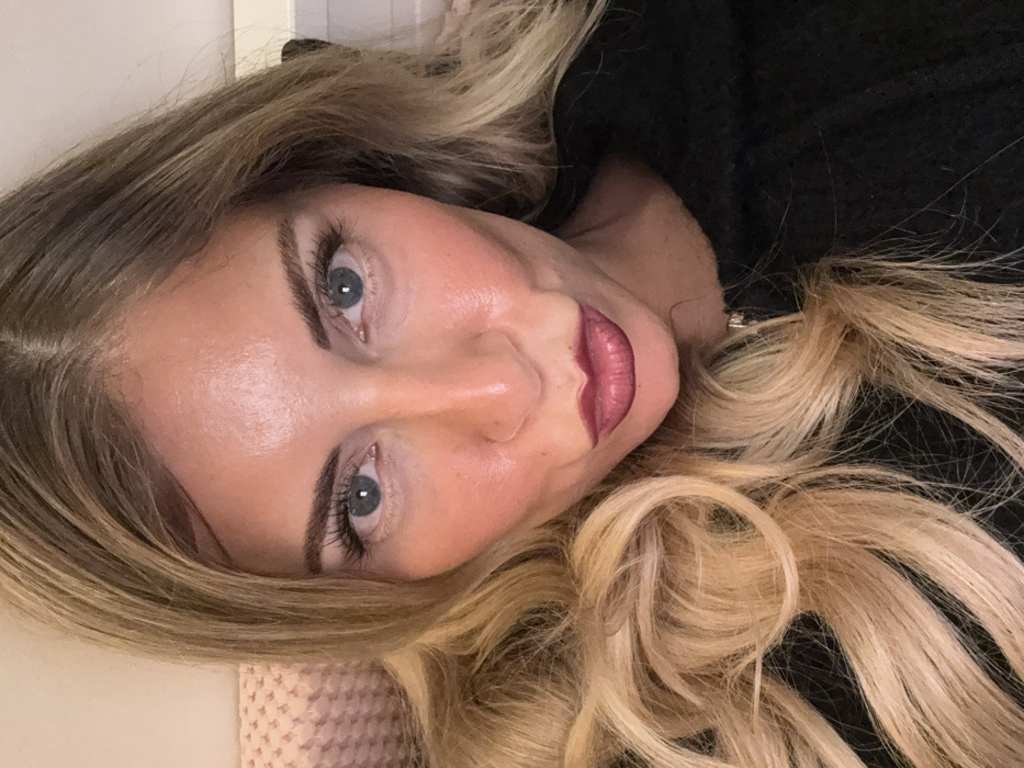 A fair-skinned woman with blonde highlighted hair and blue eyes gazes at the viewer. Her makeup includes defined eyebrows, mascara, blush, and dark red lipstick. She is wearing a black top with a visible collar. The image is rotated, showing her face at an angle. Her hair is styled in loose waves.