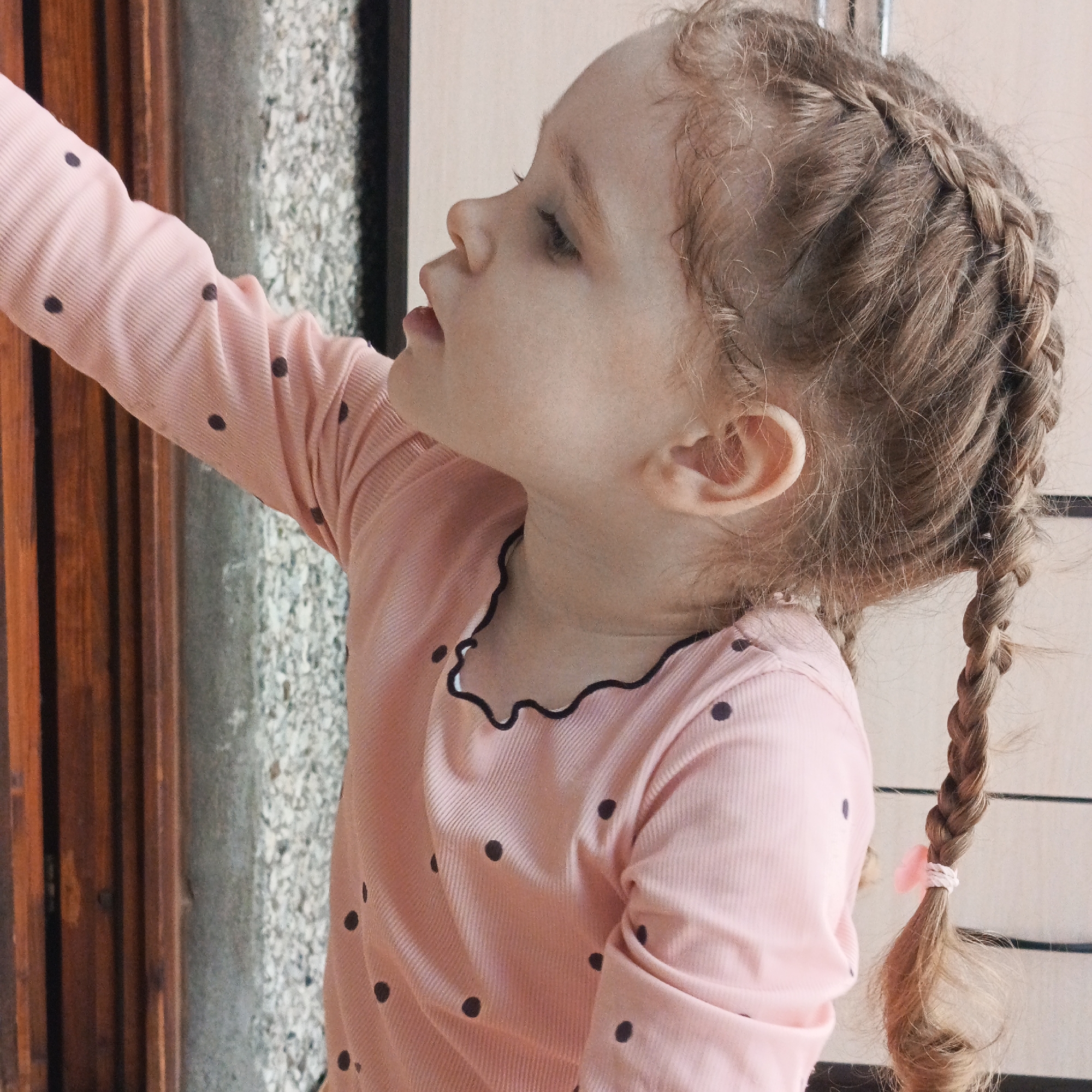 A young child with blonde hair styled in two braids, secured with pink bands, is extending their left arm towards an object outside the frame. The child is wearing a long-sleeved pink shirt with small, dark polka dots and a black wavy-edged collar. The child is standing near what appears to be a doorway, with wood paneling and a textured, light-colored wall visible. The scene is located in Марковский, Russia.