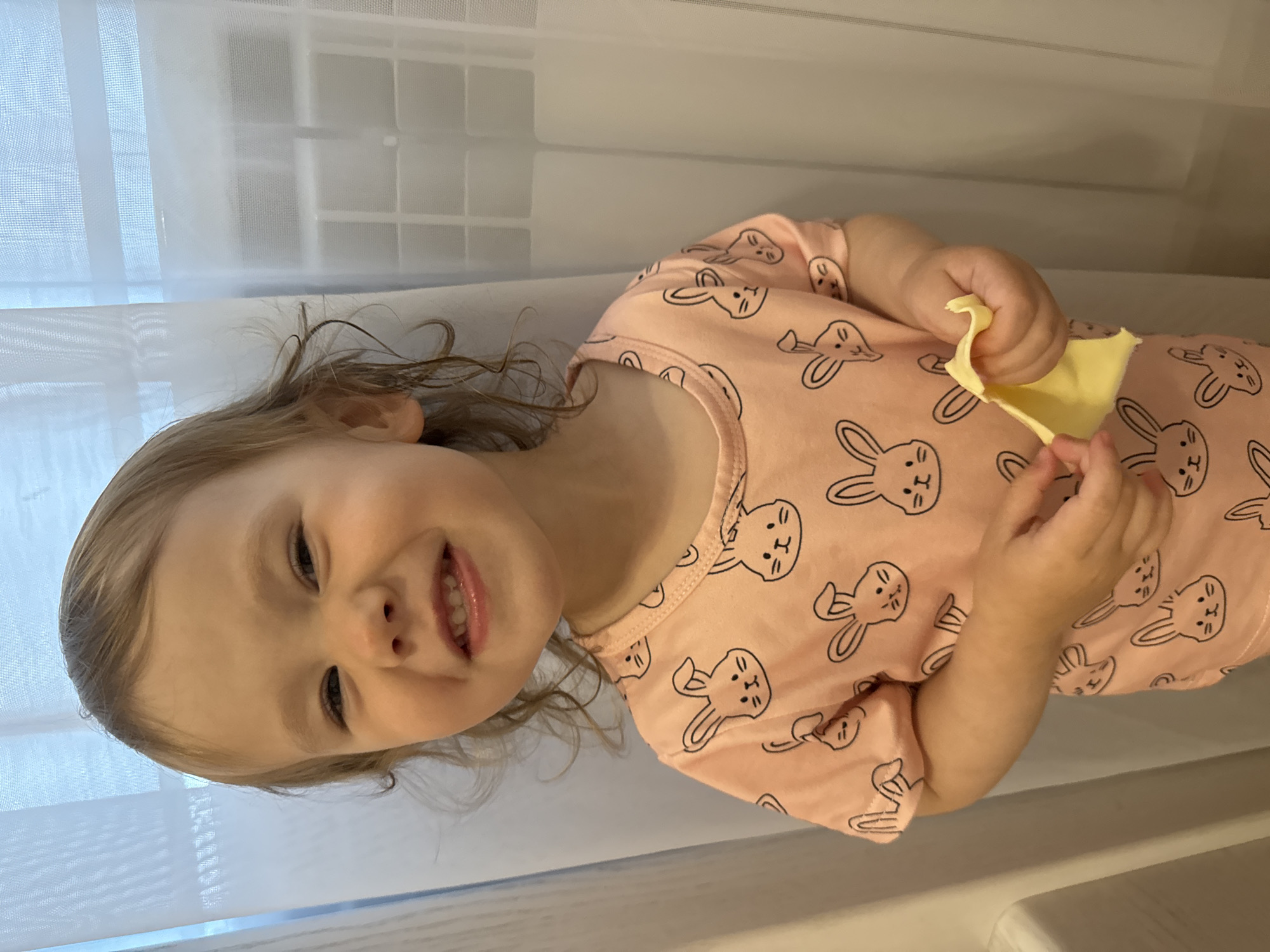 A young Caucasian child, estimated to be between two and three years old, is pictured indoors, smiling and holding a piece of yellow fabric. The child has light brown hair with curly ends and is wearing a pink t-shirt adorned with a pattern of black bunny faces. The background consists of sheer white curtains, suggesting a window or doorway. The lighting is soft and diffused. The child's expression is one of amusement or joy, with eyes crinkled at the corners and teeth visible. The yellow fabric appears to be crinkled and slightly torn at the edges.