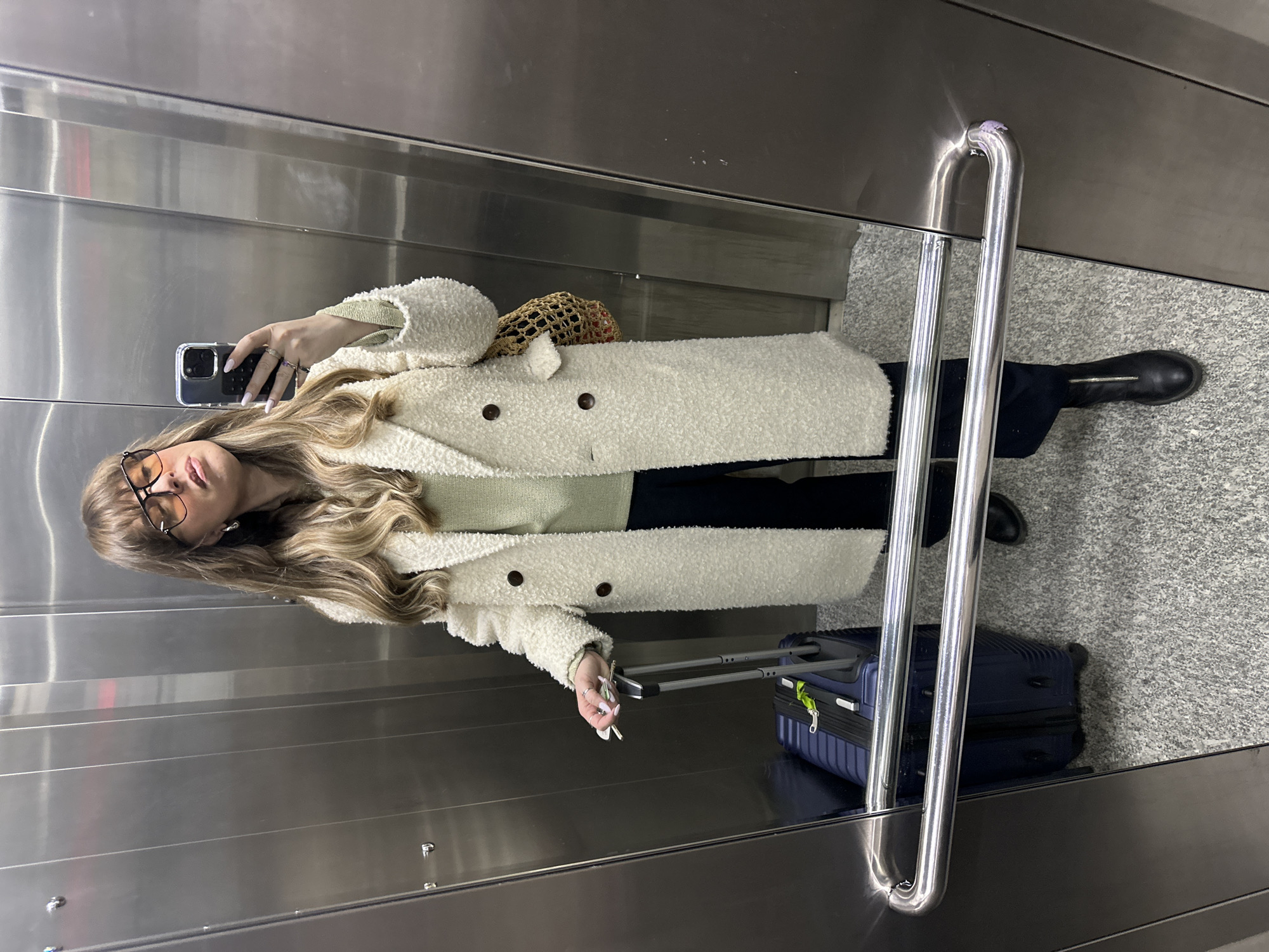 Inside a metallic elevator, a person is taking a selfie. The person is wearing glasses, a white textured coat, and dark pants, and black boots. They are holding a smartphone in their left hand. To their right is a blue suitcase. A gray patterned floor is visible. The elevator appears to be in a modern building. The location is indeterminate but presumed to be in the city of Tbilisi, Georgia.