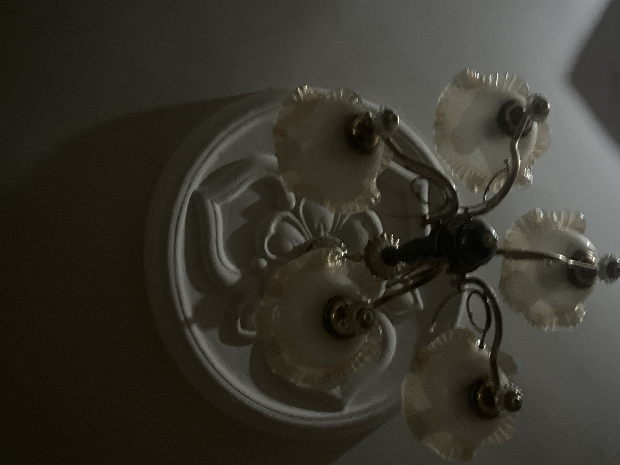 A close-up, low-angle shot captures a vintage-style chandelier hanging from a decorative ceiling medallion in what appears to be an indoor setting. The chandelier has a dark central body with multiple arms extending outwards, each supporting a ruffled, white or cream-colored lampshade. The lampshades are translucent, and the metallic accents on the chandelier, such as the bulb holders and arm connections, are visible. The ceiling medallion is ornate, with raised floral and swirling patterns painted white. The lighting in the image is dim, with shadows cast on the ceiling, suggesting it might be nighttime or an indoor space with limited illumination. The location is Jalingo, Nigeria, though this is a general detail and not visually verifiable from the image itself. There are no people or animals present in the frame. The scene is static and appears to be a still photograph of home decor.
