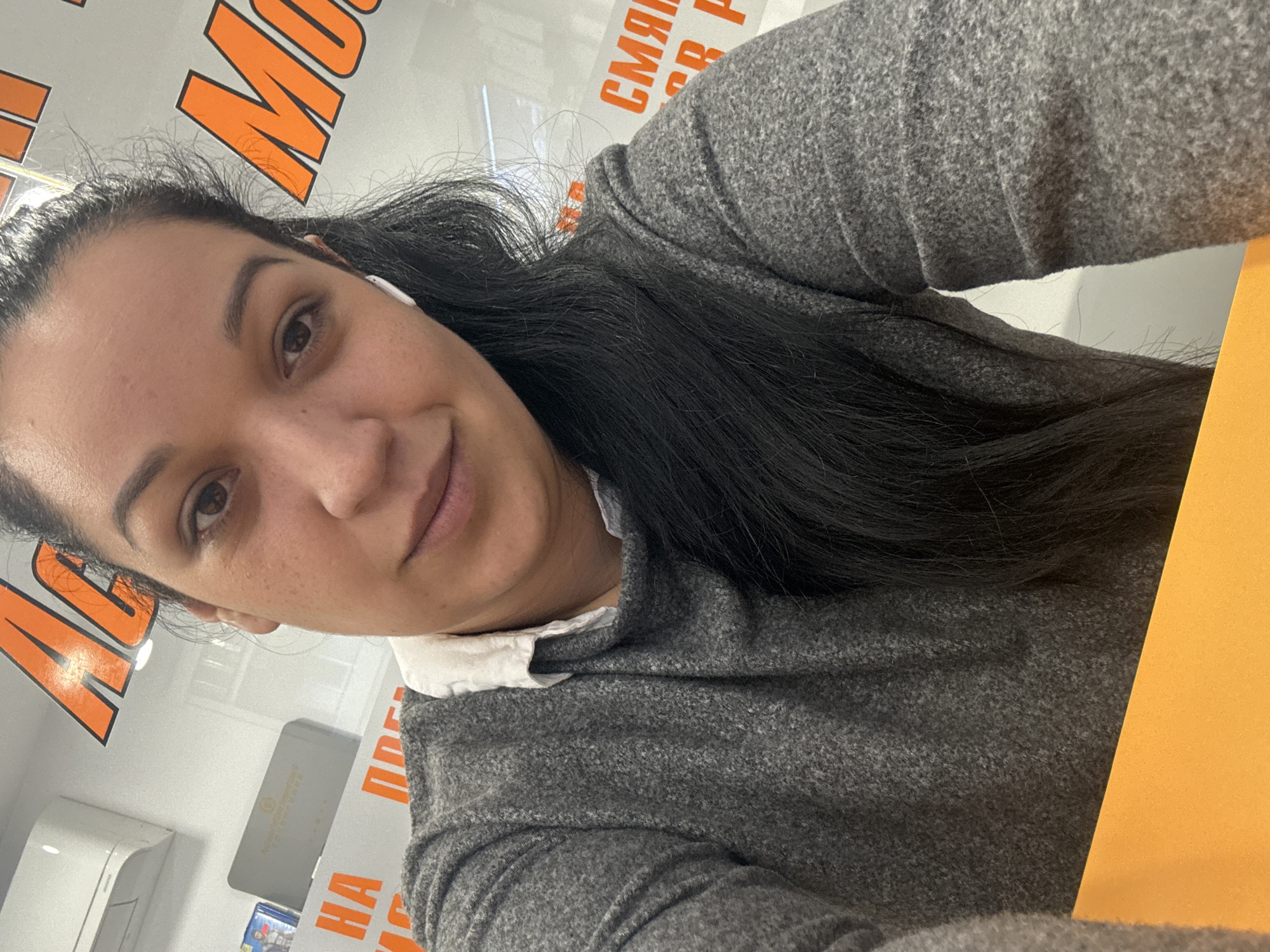 A medium close-up selfie of a woman with dark hair and brown eyes. She wears a gray wool sweater with a white collared shirt underneath. An earbud is visible in her left ear. The photo is taken indoors, with a bright orange sign with Cyrillic letters in the background, which reads "МОБИЛЕН СВЯТ Р. Петков." To the left, more signage is partially visible. A sheet of orange-colored paper is on a desk or surface on the right. The location is likely in Burgas, Bulgaria, based on the Cyrillic signage and the presence of "Р. Петков," which may indicate the name of the store owner or business establishment.