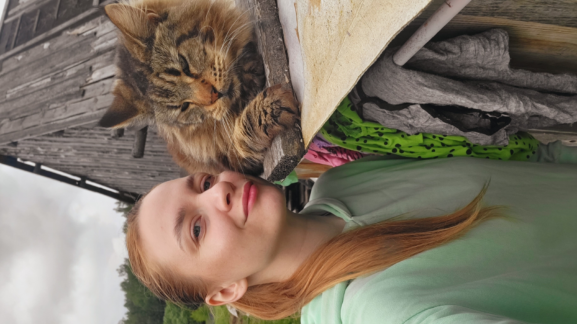 This full-length portrait, taken in Зипуново, Russia, depicts a woman and a cat positioned outdoors against a rustic wooden structure on an overcast day.
The woman occupies the left half of the frame, facing slightly right and looking directly at the viewer with a subtle smile. She has light skin, blue eyes, and light red hair pulled back into a low ponytail, with some strands falling over her left shoulder. She is wearing a light mint green hooded sweatshirt. Her head is tilted slightly toward the cat.
A large, fluffy brown and black tabby cat with amber eyes is positioned to the right of the woman, resting on a weathered wooden plank. The cat appears calm, with its eyes partially closed, and one front paw is visible, resting on the plank. Its fur exhibits a marbled tabby pattern with hints of orange-brown tones. The cat's face is close to the woman's face, suggesting a close interaction, possibly a selfie.
Behind the subjects, a tall, weathered wooden building, likely an outbuilding or barn, dominates the upper right background. Its wooden planks are dark and vertically aligned, indicating age. Above the structure, a pale, cloudy sky is visible. To the far left, a dense line of green trees suggests a natural, possibly forested, area in the distance.
Below the wooden plank where the cat rests, various items of clothing or fabric are visible, hanging or draped. These include a bright lime green fabric with black polka dots on the left, and several pieces of grey and black fabric, possibly laundry, on the right. A white metal bar or pipe is also visible horizontally in the lower right, partially obscured by the fabrics. The overall scene suggests a rural or semi-rural outdoor setting.