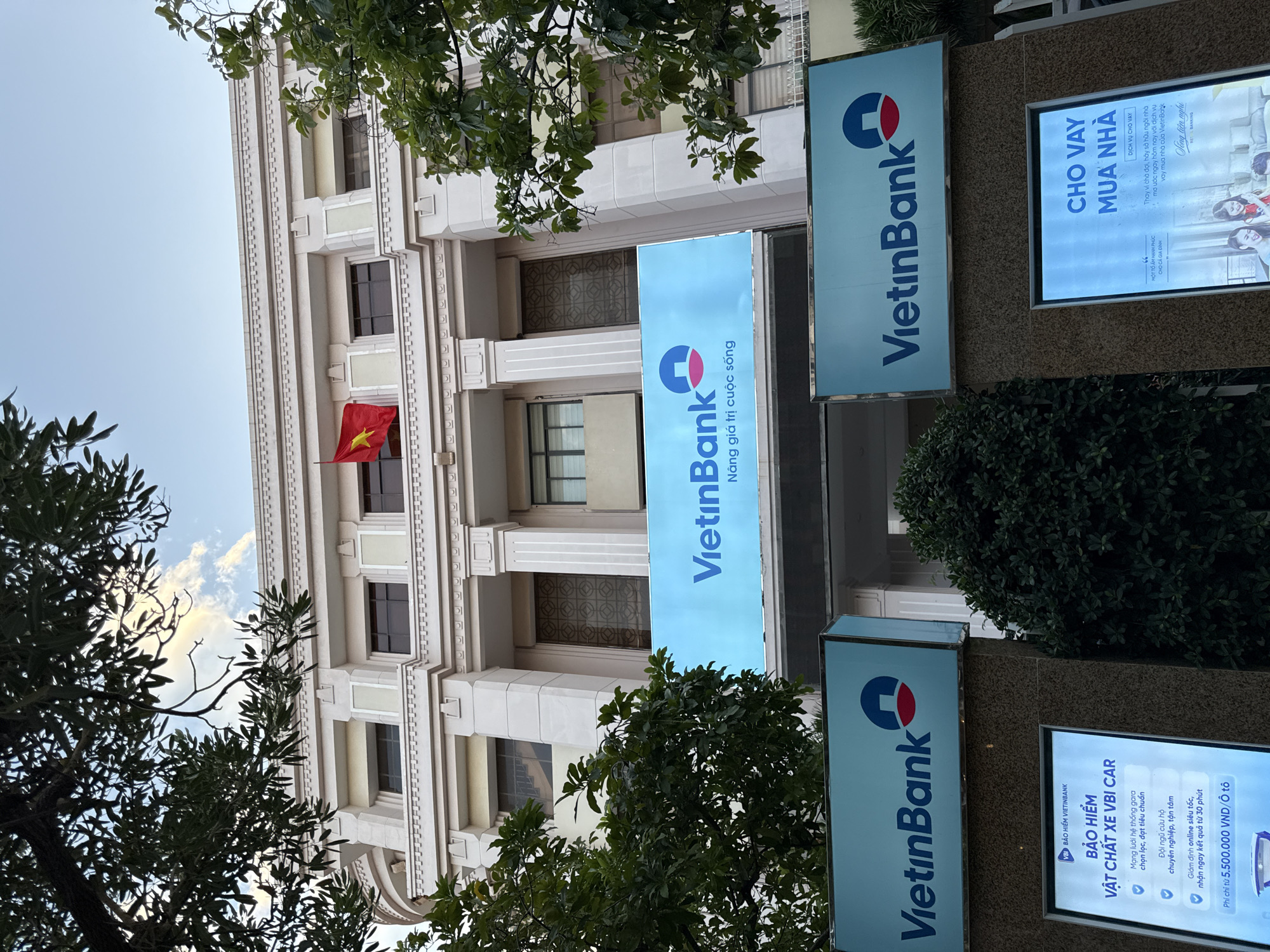 A wide-angle, low-angle shot captures the facade of a building in Nha Trang, Vietnam, dominated by a large, light blue sign displaying "VietinBank" with its logo. A Vietnamese flag, red with a yellow star, flutters from a pole on the building's upper section. Lush green trees partially obscure the building's architecture, which features cream-colored stone and classical columns. Below the main sign, two smaller, illuminated display boards, also featuring the VietinBank logo, are mounted on pillars. The left display board advertises "Bảo Hiểm Vật Chất Xe VBI CAR," detailing benefits and a price of 5,500,000 VND. The right display board promotes "Cho Vay Mua Nhà" (Home Loans), showcasing families and the VietinBank brand. The sky above is a clear, pale blue, with some wispy clouds. The overall impression is of a modern financial institution in an urban setting.