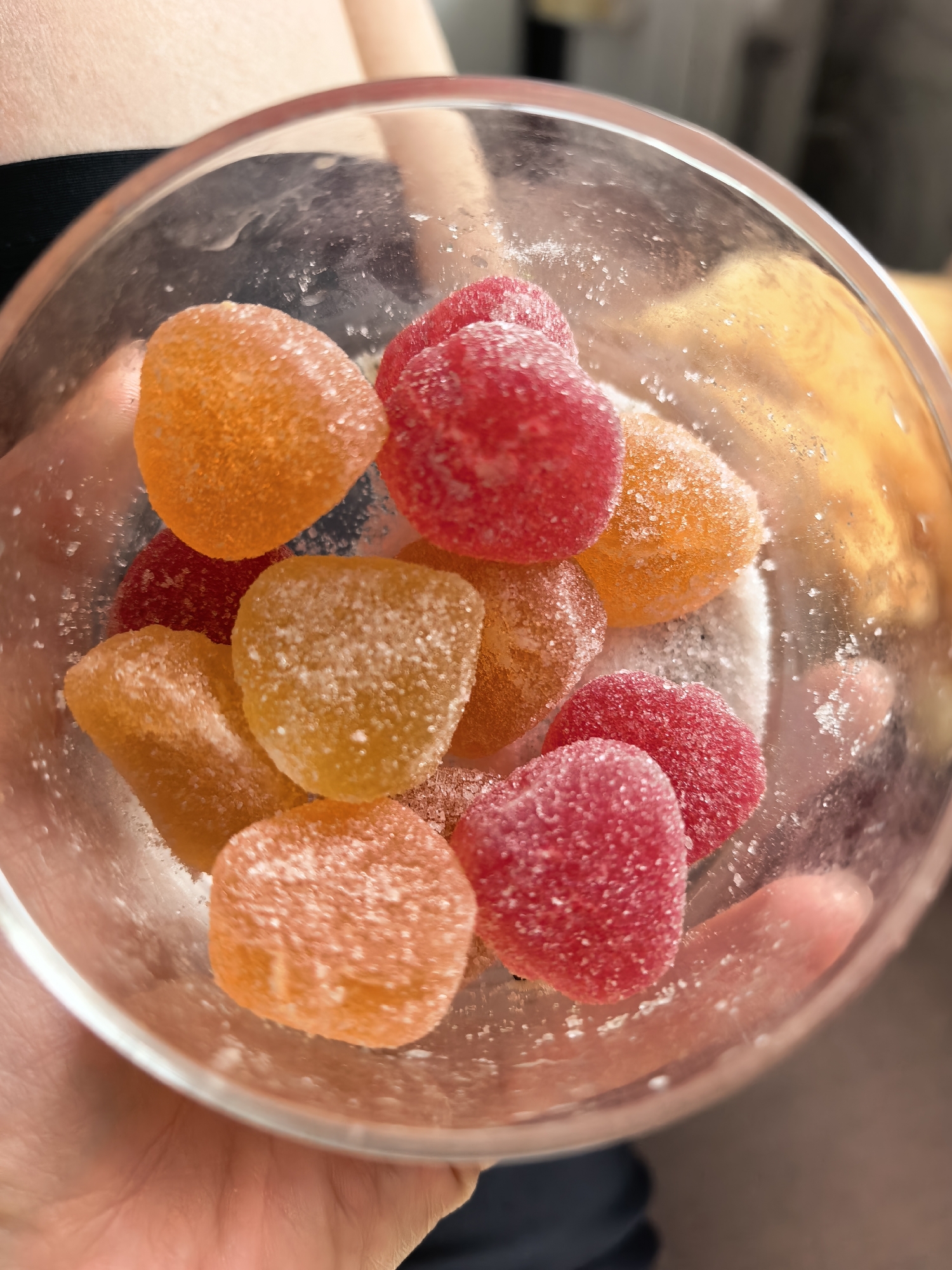A close-up, overhead shot shows a clear glass bowl filled with heart-shaped gummy candies. The candies are coated in sugar and are a mix of orange and red colors. The bowl is held by a person's hands, and the background is blurred. The image does not contain any information that can be used to determine its location in Sterlitamak, Russia.