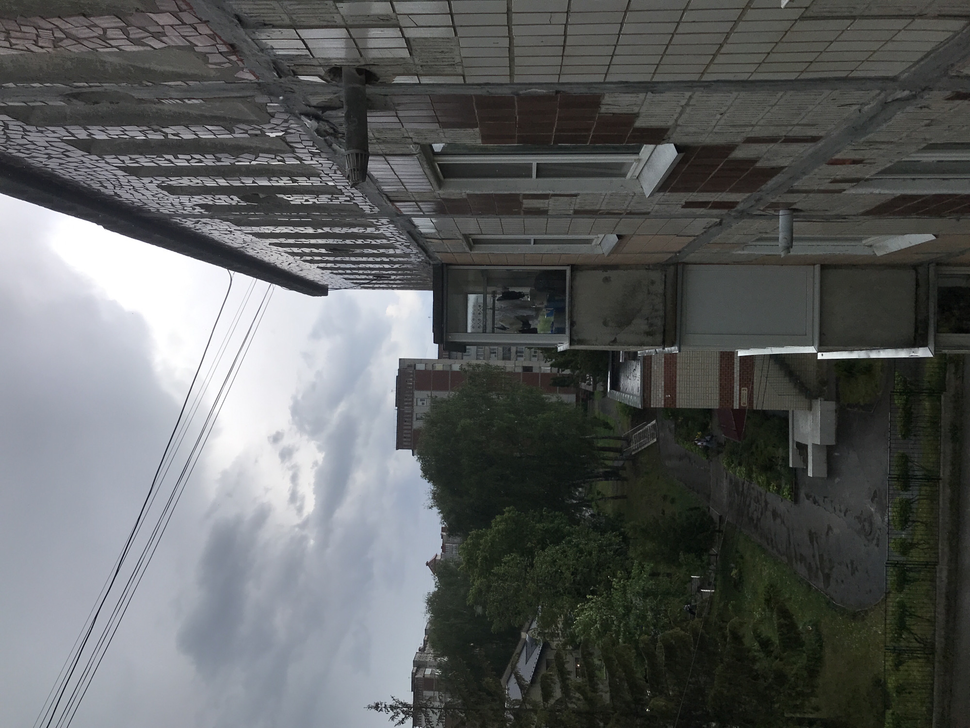 The image depicts an overcast day in what appears to be a residential area. In the foreground is part of a building exterior, showing walls with tiled surfaces and windows, some of which are partially open or have extensions. Further in the background, trees line a pathway or street, obscuring parts of taller apartment buildings. The presence of dark, heavy clouds suggests recent or impending rain. Based on the location, the scene depicts a typical Soviet-era residential area in Ternopil, Ukraine.