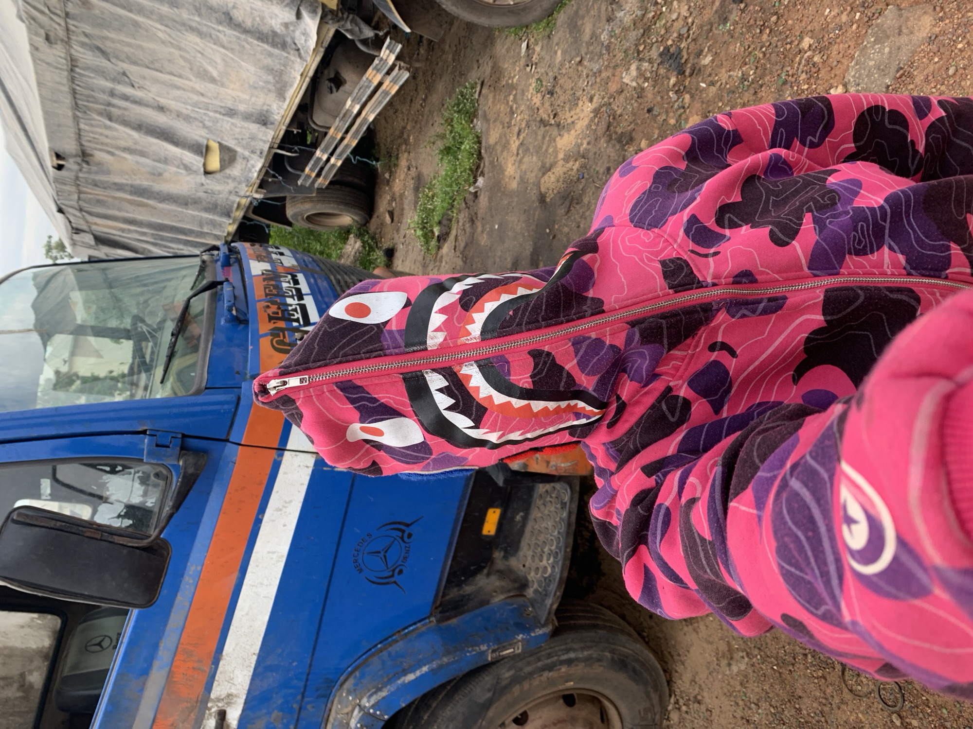 The image depicts an outdoor scene in Rafin Par, Nigeria, featuring a person taking a selfie in the foreground, positioned in front of two heavy-duty trucks.
In the immediate foreground, a person is visible from the chest up, holding a camera or smartphone to take a selfie, with their face fully obscured by the hood of their attire. They are wearing a full-zip hoodie in a hot pink and dark purple camouflage pattern. The hood features a distinctive "shark face" design, with prominent white teeth outlined in black, red gums, large white eyes with black pupils, and a red circular accent on the forehead. The zipper of the hoodie is silver-colored and runs up the center.
Behind the person, to the left, is the front and side of a large, blue Mercedes-Benz cab-over-engine truck. The truck's blue paint is faded and dirty, with visible chipping. It features two horizontal stripes, orange over white, across its side panel. A black "MERCEDES BENZ" logo, incorporating the three-pointed star, is stenciled or stickered onto the lower part of the blue fender above the front left wheel. Faint, worn white lettering, possibly "CAPTAIN," is visible above the orange stripe on the cab door. The front left tire and part of the muddy running board are also visible.
To the right and further back, partially obscured by the blue truck and the person, is another large truck. This truck's cargo area is covered by a heavily wrinkled, dirty, light grey canvas tarp, showing several tears, including a rectangular one on its side. The rear wheels of this truck are visible on the far right.
The ground throughout the scene is composed of reddish-brown dirt and mud, with sparse patches of green weeds. The sky above is overcast, indicating cloudy conditions.