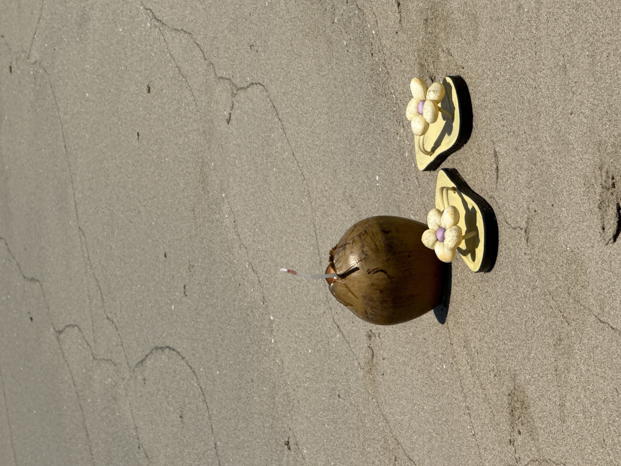 The image depicts a coconut with a straw inserted, lying on a sandy surface, alongside a pair of yellow sandals with flower decorations. The location appears to be a beach, possibly in Đà Nẵng, Vietnam, given the likelihood of coconuts and beach sandals in such a coastal city. There are no people visible. The primary objects are the coconut, straw, and sandals. No actions or interactions are evident in this static image. The environment is characterized by light-colored sand.