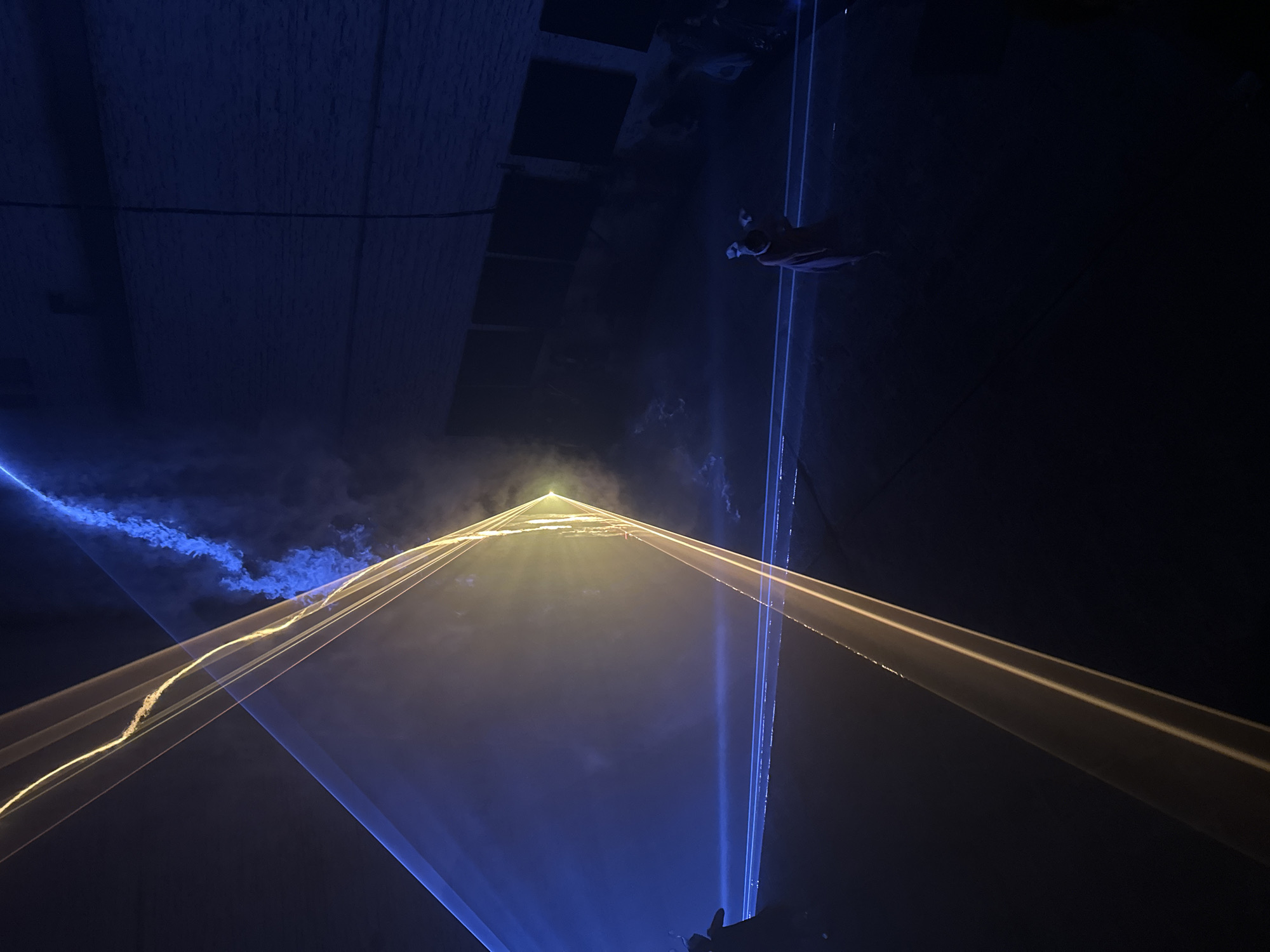 The image presents a scene in what appears to be a large indoor venue, possibly located in Berlin, Germany given the style of architecture. The primary visual feature is a display of laser lights. The beams are a mix of yellow and blue, creating geometric patterns in the dark space. The beams converge, forming a triangle. The light illuminates suspended particulates in the air.
In the background, there is a visible architectural element that appears to be part of the building's structure. An additional figure is seen in the very top right of the image, possibly suspended in the air. The overall atmosphere is dimly lit, emphasizing the intensity and clarity of the laser light show.