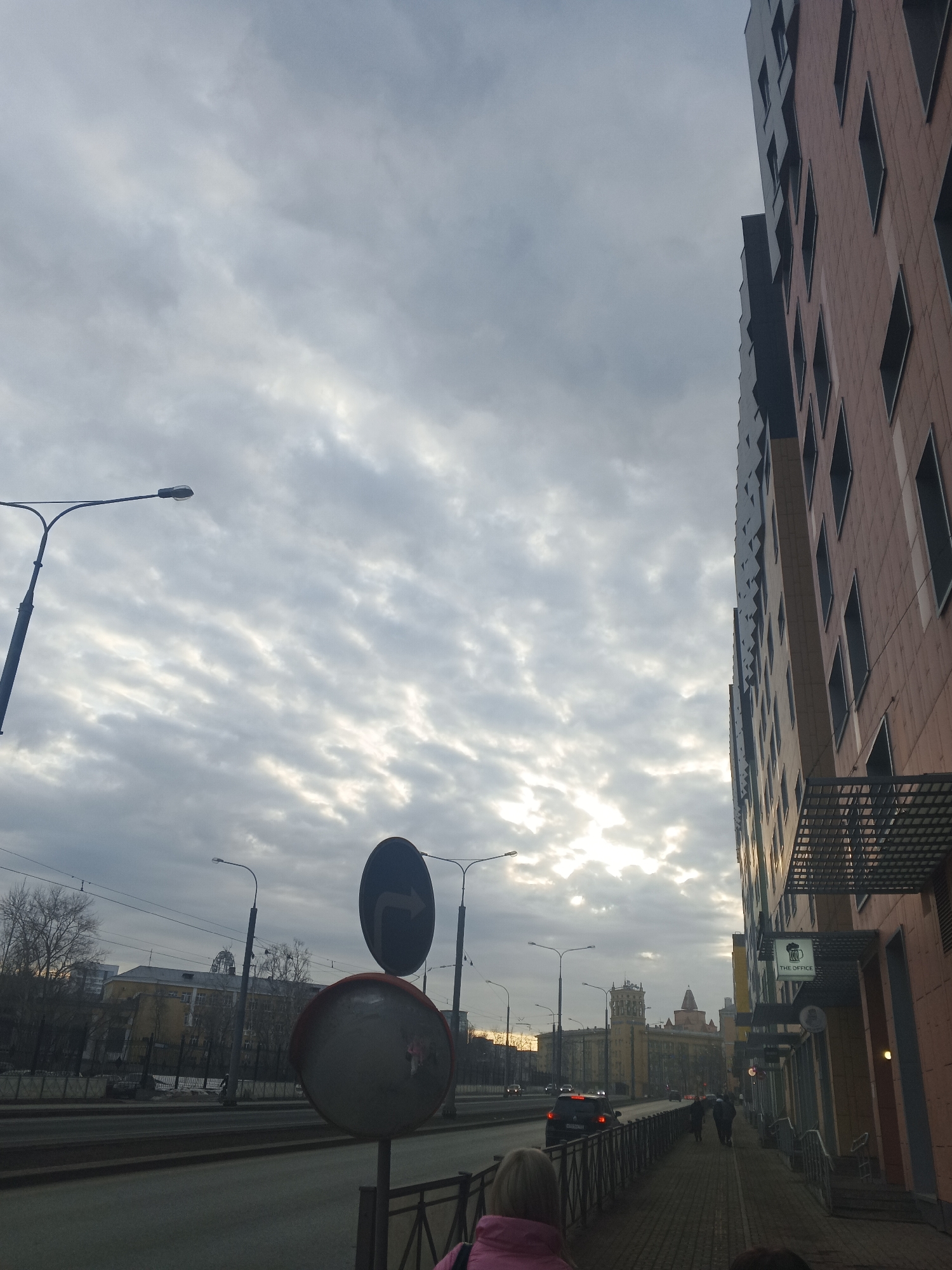 An eye-level outdoor shot on a cloudy day in Perm, Russia depicts a street scene. On the right side of the image is a multi-story building with a peach facade and dark window accents. A sign reading "THE OFFICE" is visible.
A pedestrian walkway, defined by a metal railing, runs along the building's edge. Several people walk in the distance.
To the left is a street with multiple lanes. A car drives along the road. Traffic signs are visible, including one showing a curved arrow. A reflective safety mirror is mounted below.
Across the street is a building with a yellow facade. A tower-like structure rises in the center. Bare trees are scattered in the background. Street lamps line the street.
The sky is filled with striated grey clouds. Sunlight filters through a gap.