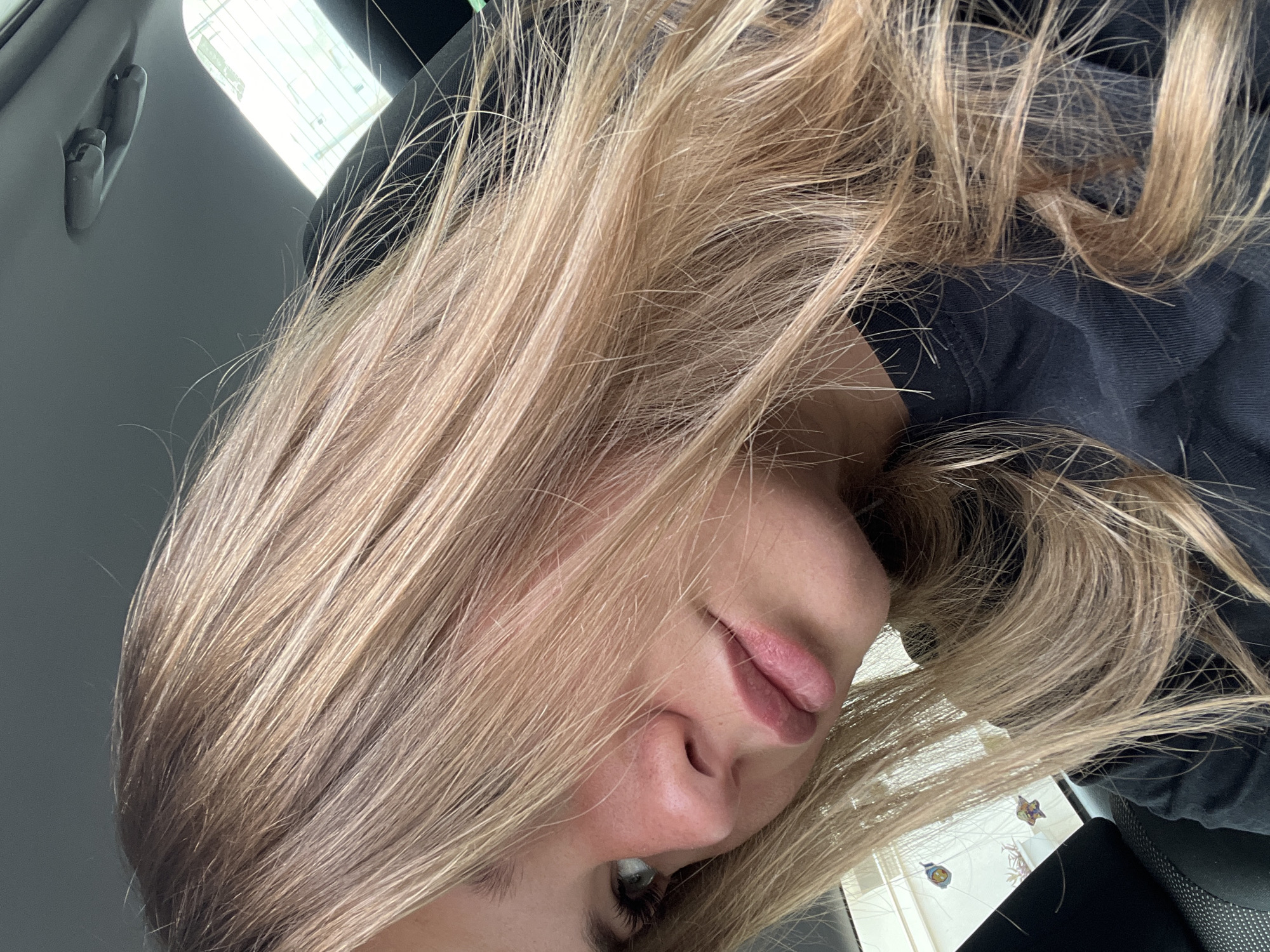 The image depicts a person with blonde hair inside a car. The person is positioned such that the camera is looking up at them. The car is likely a private vehicle. The location in Budva, Montenegro is unknown.