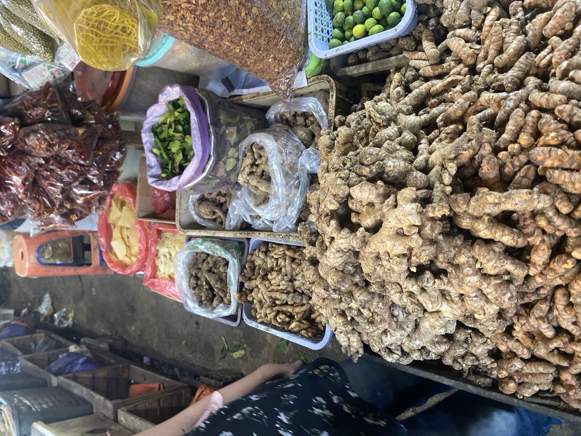 The image shows a market stall in Cikupa, Indonesia, with a focus on various produce and dried goods. In the foreground, a large pile of ginger and turmeric roots is prominently displayed in a black plastic container. To the right, a light blue plastic basket holds a collection of small green limes. Behind these, several plastic bags contain other items: one large bag is filled with what appears to be dried chili peppers, and another contains a granular brown substance, possibly dried spices or ground nuts. Smaller plastic bags hold darker, gnarled roots, possibly more ginger or similar rhizomes. A purple-trimmed bag contains chopped green leaves, likely herbs. Further back, a red plastic basket holds dried, yellowish chips, and another red container holds a similar item. A red plastic stool is visible in the upper background, alongside stacked shelves containing various items. A person's arm and shoulder, wearing a black and white patterned garment, are visible on the left side of the image, partially obscuring the stall. The ground appears to be a concrete or dirt surface, typical of an outdoor or semi-enclosed market.