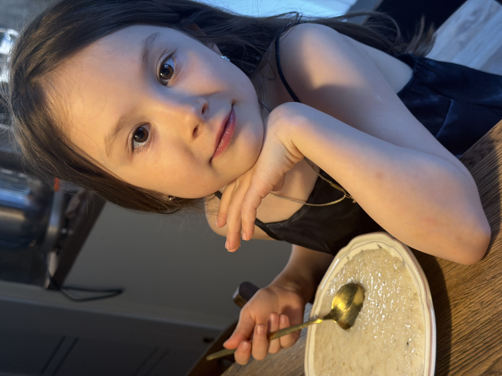 A close-up, angled portrait features a young girl, approximately 5-8 years old, looking directly at the camera with a gentle, slight smile. She has long, dark brown hair, fair skin, and large, expressive dark brown eyes. Her head is tilted, and her left hand is resting casually under her chin. She wears a dark, possibly black, camisole or dress with thin straps, accessorized with small earrings and a delicate gold-colored necklace.
In her right hand, she holds a golden spoon, which is positioned in an oval-shaped white bowl. The bowl contains a light-colored, creamy substance, likely oatmeal or porridge, suggesting she is in the process of eating or about to begin a meal. The bowl and her arm rest on a wooden table surface.
The background is softly blurred, showing dark, indistinct shapes of furniture or walls, with some metallic objects visible to the far left. The lighting is soft and warm, casting highlights on her hair and shoulders, creating an intimate and cozy indoor atmosphere. The scene captures a moment of quiet engagement within a domestic setting. There is no visible text in the image.