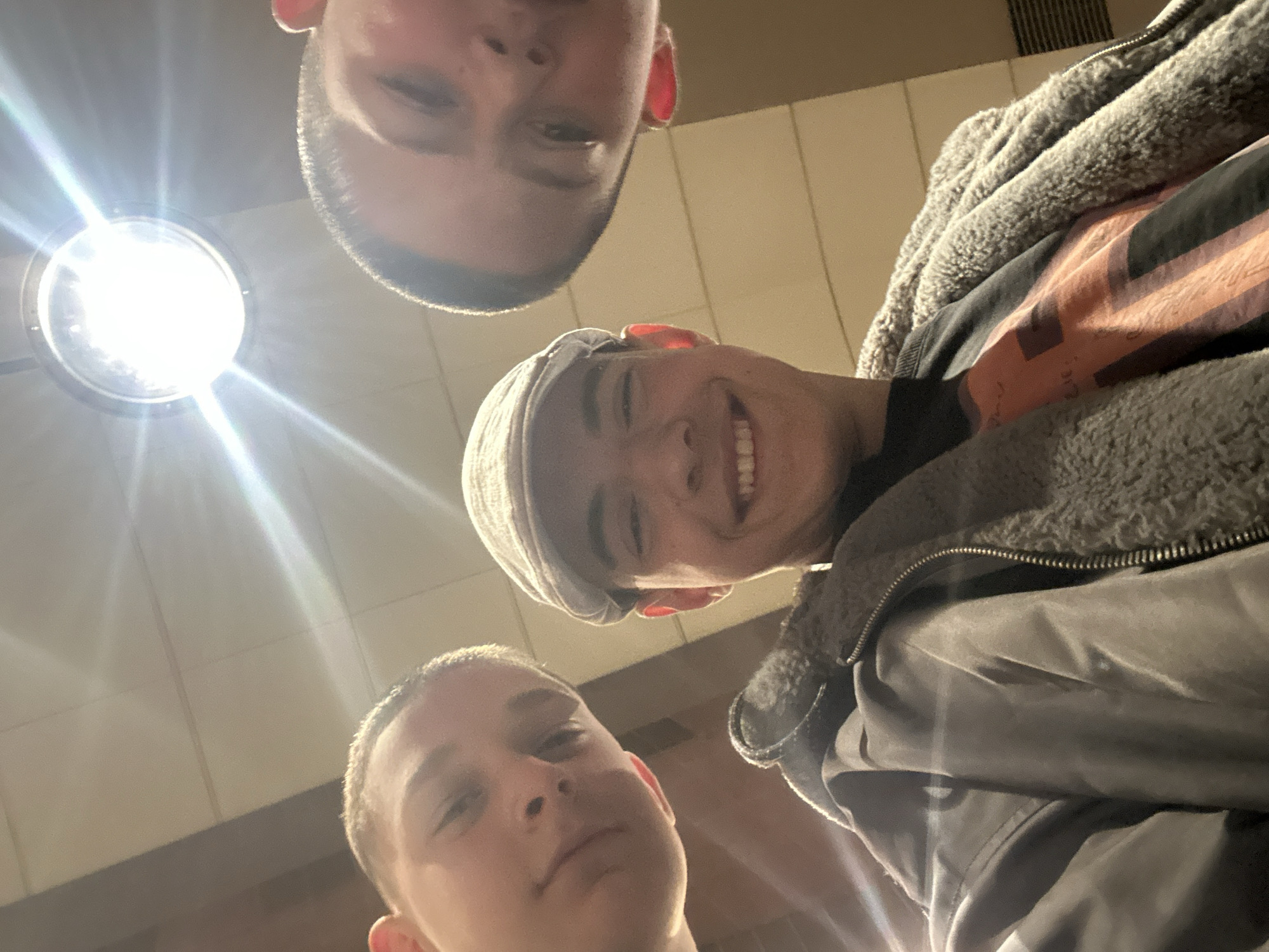 The image shows three young men taking a selfie from a low angle, looking upwards. One is wearing a cap, and another a jacket with a fuzzy lining. There is a bright light fixture in the background. Based on the architectural style of the wall visible, and the presence of what appears to be a ceiling vent, the location may be an indoor public space. This image does not contain enough details to determine if the picture was taken in the city of Sofia, Bulgaria.