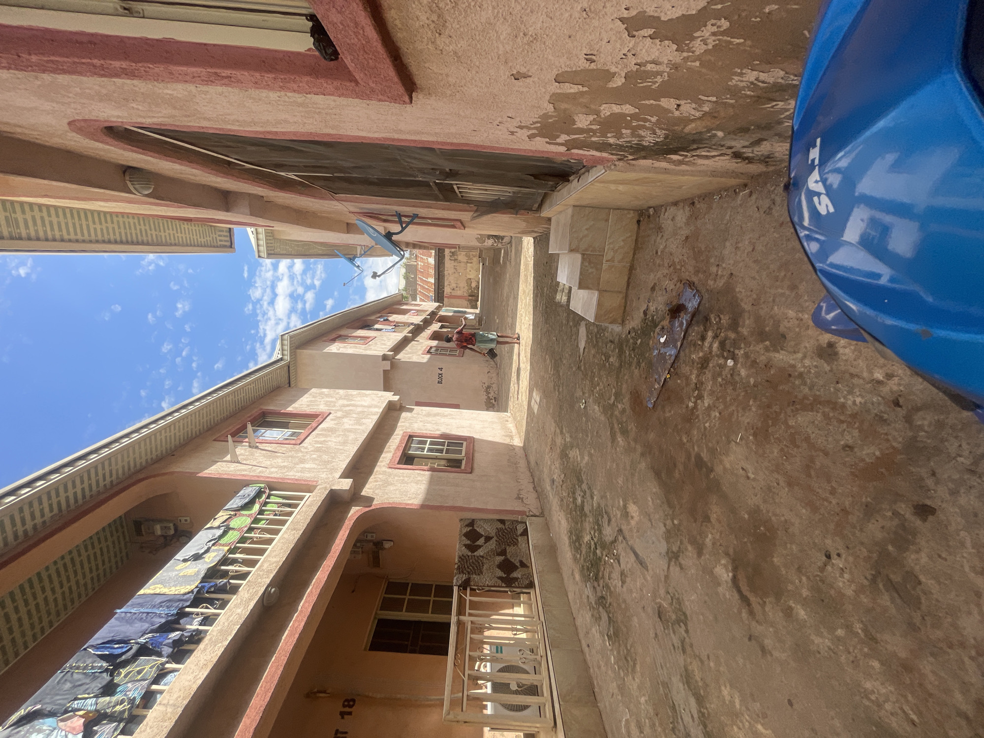 The image displays a narrow outdoor passageway or alleyway between two multi-story residential buildings, presented from a low, tilted perspective. The scene is illuminated by bright daylight under a clear blue sky with scattered white clouds, indicating a sunny day. The location is Lokoja, Nigeria.
The buildings are light-colored, possibly beige or pale pink, with reddish-brown accents around windows and balconies. Signs of wear, such as peeling paint and discoloration, are visible on the walls. On the left side, a balcony on one of the buildings features various items of clothing, including shirts and trousers, hung on a rack to dry. An outdoor air conditioning unit is also present on a lower balcony. On the right building, two small blue satellite dishes are mounted on the exterior wall.
In the middle distance, a person, possibly a child or young adult, stands in the alleyway wearing a patterned shirt and shorts, with their arms slightly outstretched. On the far right, part of a blue motorcycle or scooter is visible, with the brand name "TVS" clearly printed in white on its side. A set of concrete steps leads upwards on the right side of the alleyway.
The ground surface is unpaved, appearing to be dirt or worn concrete, with some debris and patches of discoloration. Visible text includes "TVS" on the motorcycle, "BLOCK 4" painted on the wall of one building, and "AT 18" inscribed near a lower balcony railing.