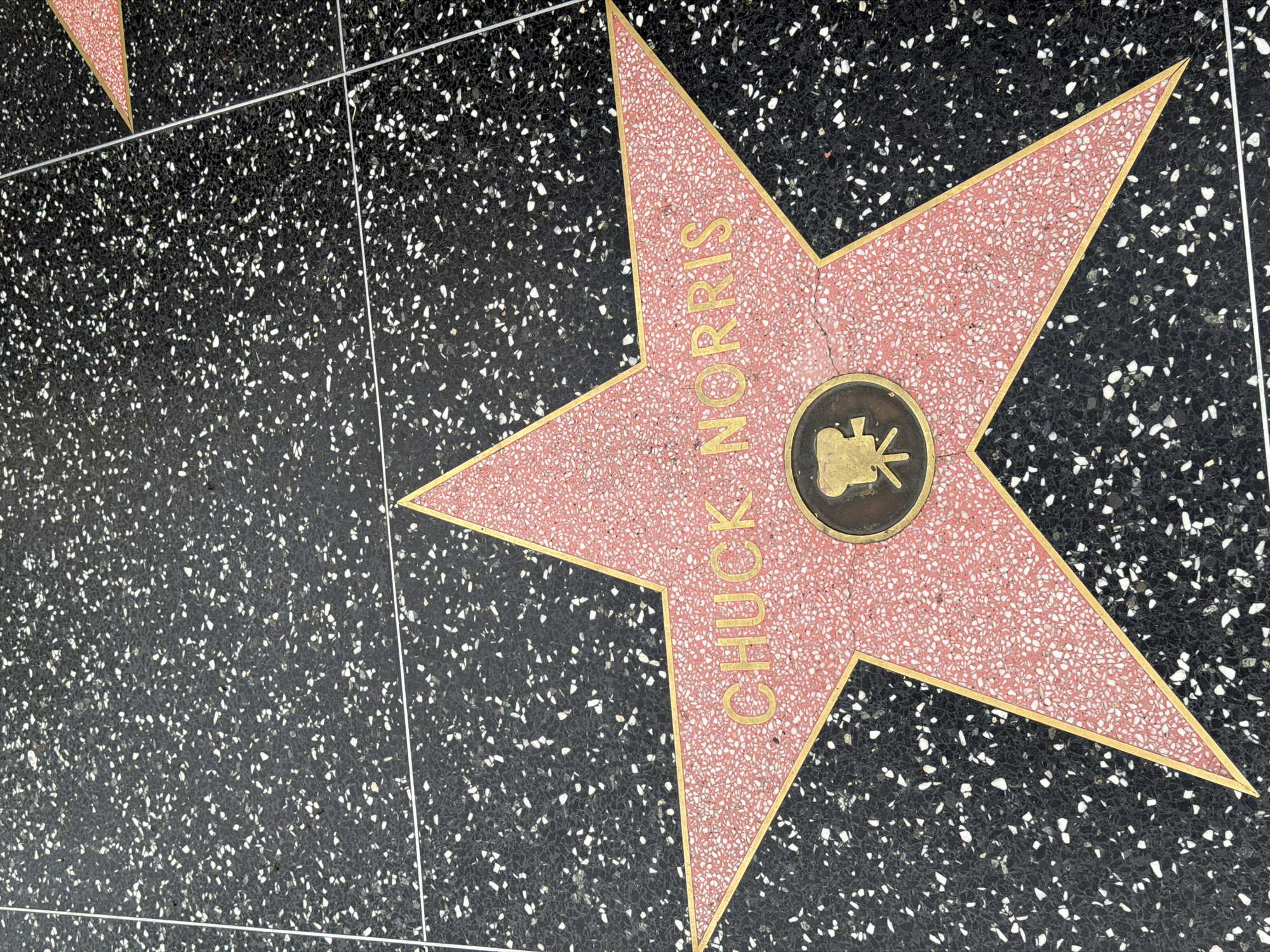 The image shows Chuck Norris' star on the Hollywood Walk of Fame in Los Angeles, California. The star is pink with gold lettering and a gold outline. In the center is a bronze medallion with a film camera symbol. The surrounding pavement is black with small white speckled aggregate. A small portion of another star is visible in the upper left corner. White dividing strips are also present.