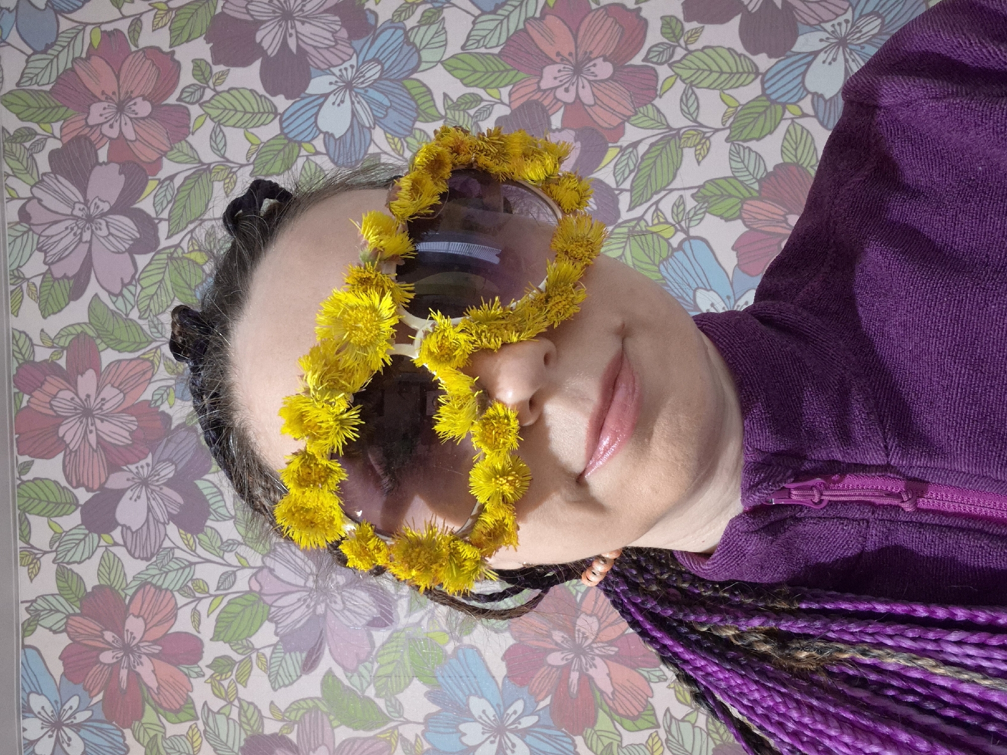 A rotated close-up portrait captures a person looking directly at the camera with a slight, pleasant smile. The individual has dark hair styled with long, multi-toned purple and brown braids visible on the right side of the frame. They are wearing large, purple-tinted sunglasses, which are extensively adorned with small, vibrant yellow flowers, possibly dandelions, around the entire frame. The person is dressed in a textured purple top, featuring a matching purple zipper on the right side. The background is a light-colored surface with a repeating floral pattern, showcasing outlined flowers in shades of pink, purple, and blue, interconnected by green leaves and stems. The setting appears to be indoors. No text is visible.