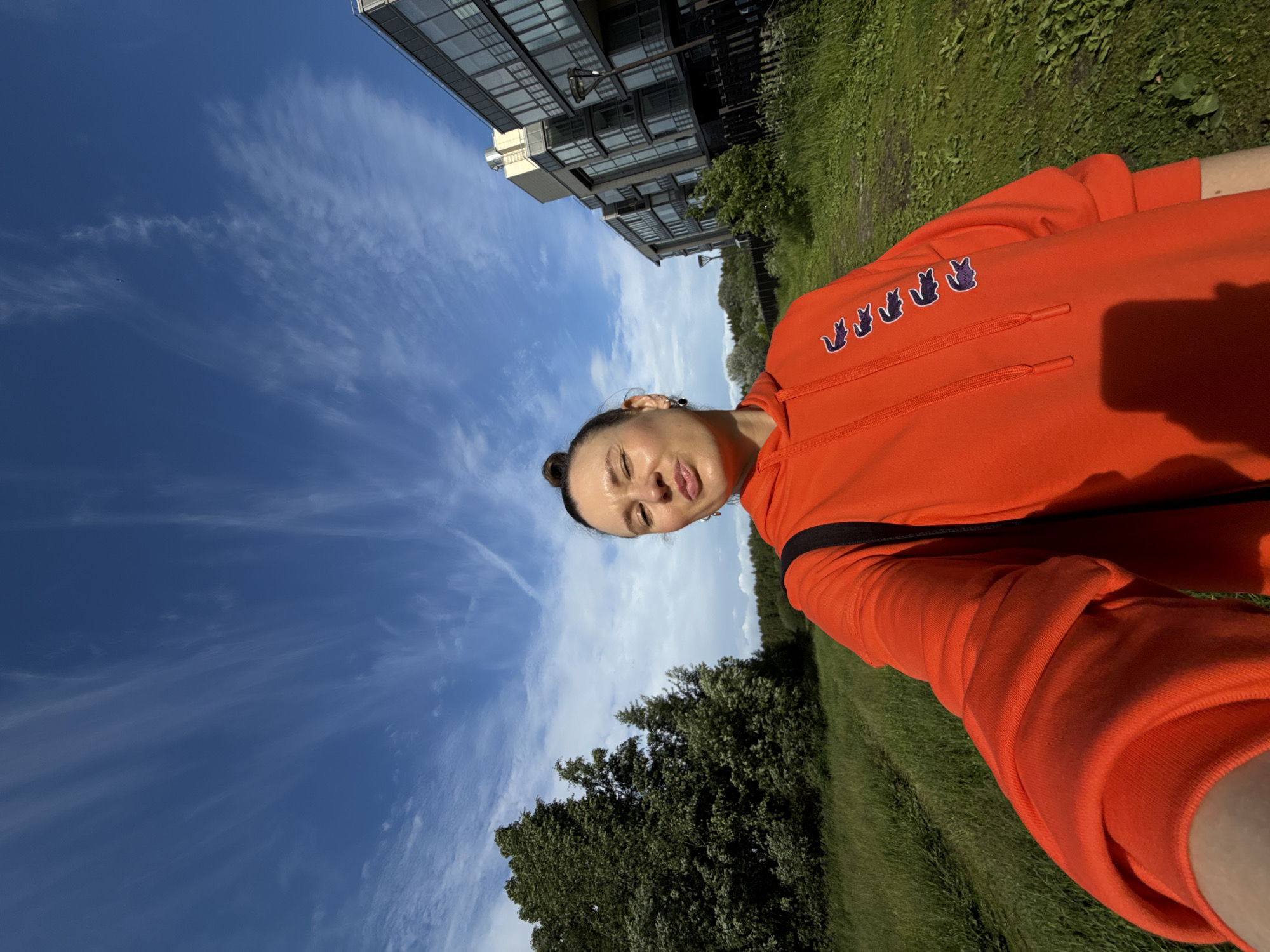 A woman is taking a selfie outdoors in front of a modern building. She is wearing an orange hooded sweatshirt with multiple crocodile logos on the left side. Her hair is pulled up into a bun, and she is making a puckered expression at the camera. The background shows a grassy area with trees and a multi-story building with large windows. The sky is blue with wispy white clouds. The image appears to have been taken on a sunny day. The architectural style of the building, characterized by its glass facade and modern design, suggests a contemporary urban development, possibly in a suburban area of Saint Petersburg, Russia, where such residential complexes are common. The presence of greenery and open space hints at a newer neighborhood with planned landscaping, juxtaposing urban living with a connection to nature.