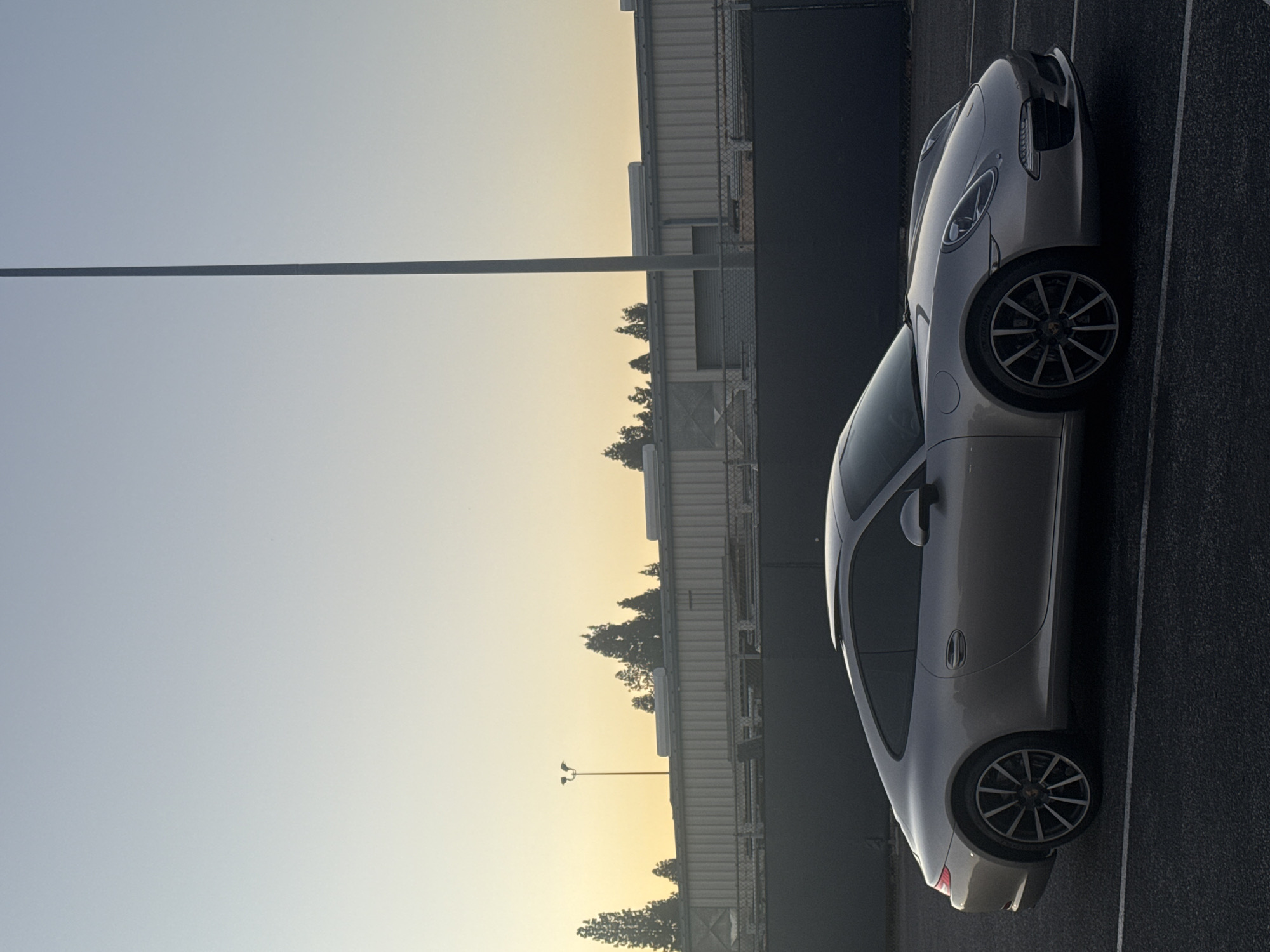 A silver Porsche 911 is parked in an asphalt parking lot at dusk. The car is angled slightly towards the viewer, with its rear quarter panel and front fender visible. The vehicle's sleek, aerodynamic design is highlighted by the soft, golden light of the setting sun.
In the background, a large, industrial-looking building with a corrugated metal facade stretches across the frame. The building appears to be a warehouse or factory, with a dark fence in front of it. Several tall, slender trees are silhouetted against the sky behind the building, their branches reaching upwards.
A tall, thin pole, possibly a lamppost, stands prominently in the middle ground, reaching high into the clear, pale blue sky. The sky itself is a gradient of light blue at the top, transitioning to a warm yellow and orange hue near the horizon, indicating the sun is setting. The overall impression is one of urban industrial scenery bathed in the serene glow of twilight. There are no people visible in the image.