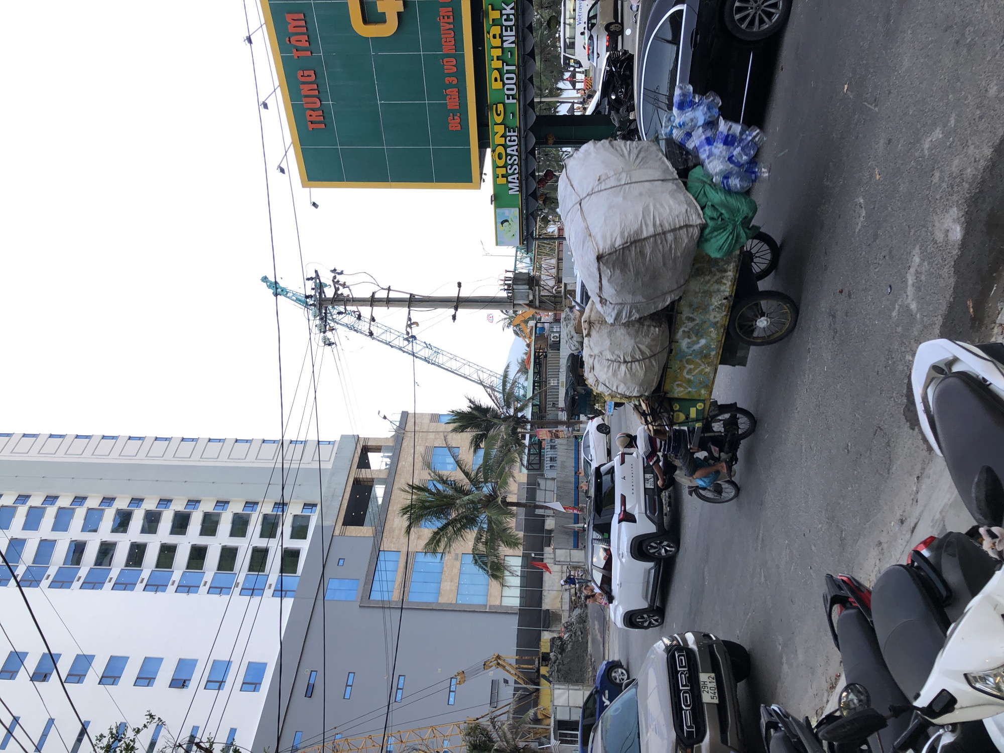 In this daylight, street-level scene in Da Nang, Vietnam, a cart laden with large, white sacks and a bundle of plastic bottles is being pulled by a man on a motorcycle. The cart, which appears to be a makeshift trailer, has two wheels and is decorated with painted yellow and blue designs. The man operating the motorcycle is wearing a striped shirt and shorts. Several other vehicles are visible, including a white SUV, a black sedan, and a brown Ford pickup truck. In the background, a tall, modern building with blue-tinted windows stands out against the sky, alongside construction cranes and scaffolding, indicating ongoing development. A large green billboard with yellow lettering is also present, advertising "TRUNG TÂM" (Center) and "HỒNG PHÁT MASSAGE - FOOT - NECK." The street is paved, and several motorcycles are parked along the side.