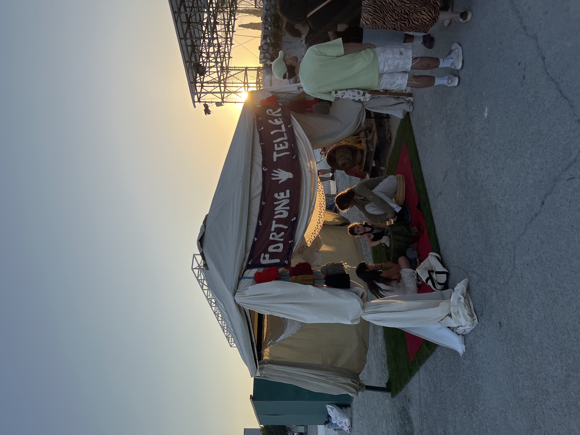 The image captures an outdoor scene at sunset in Limassol, Cyprus. A light-colored, canvas tent, featuring a prominent red banner with "FORTUNE TELLER" and white hand symbols, stands on a paved concrete surface. Artificial green grass and a red mat are laid out in front of the tent's entrance. Inside, three individuals are seated on cushions; two women appear to be engaged in a fortune-telling interaction, while a third person sits to their left. Colorful fabric tassels hang near the banner. To the right of the tent, a man in a light green t-shirt, patterned shorts, and a green baseball cap stands, facing left towards the tent, holding a white patterned tote bag. Another woman in a patterned skirt and other figures are partially visible, standing nearby. In the background, large metal scaffolding structures, consistent with event staging, are silhouetted against the setting sun, which casts a warm, orange glow through the framework. A green rectangular container is visible on the far left. The concrete ground exhibits cracks and wear.
