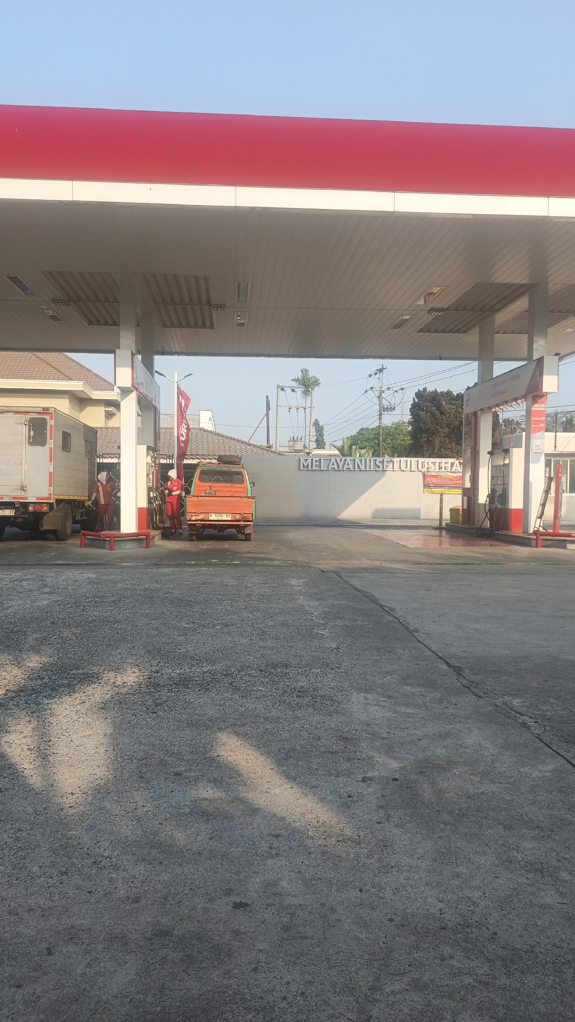 The image depicts a gas station in Rendeng, Indonesia, under a clear blue sky during daylight hours. The foreground is a concrete surface with scattered shadows. In the mid-ground, a gas station canopy with a red and white roof extends overhead. Two fuel pumps are visible, with one in use by an orange truck. Two individuals wearing red and white uniforms, likely gas station attendants, are interacting near the fuel pump. One attendant is attending to the orange truck, while another appears to be interacting with a white delivery truck parked to the left. A building with a tiled roof and white walls is partially visible behind the trucks and attendants. A sign on a white wall in the background reads "MELAYANISETULUSHA". The overall scene suggests a typical operational day at a gas station in Rendeng.
