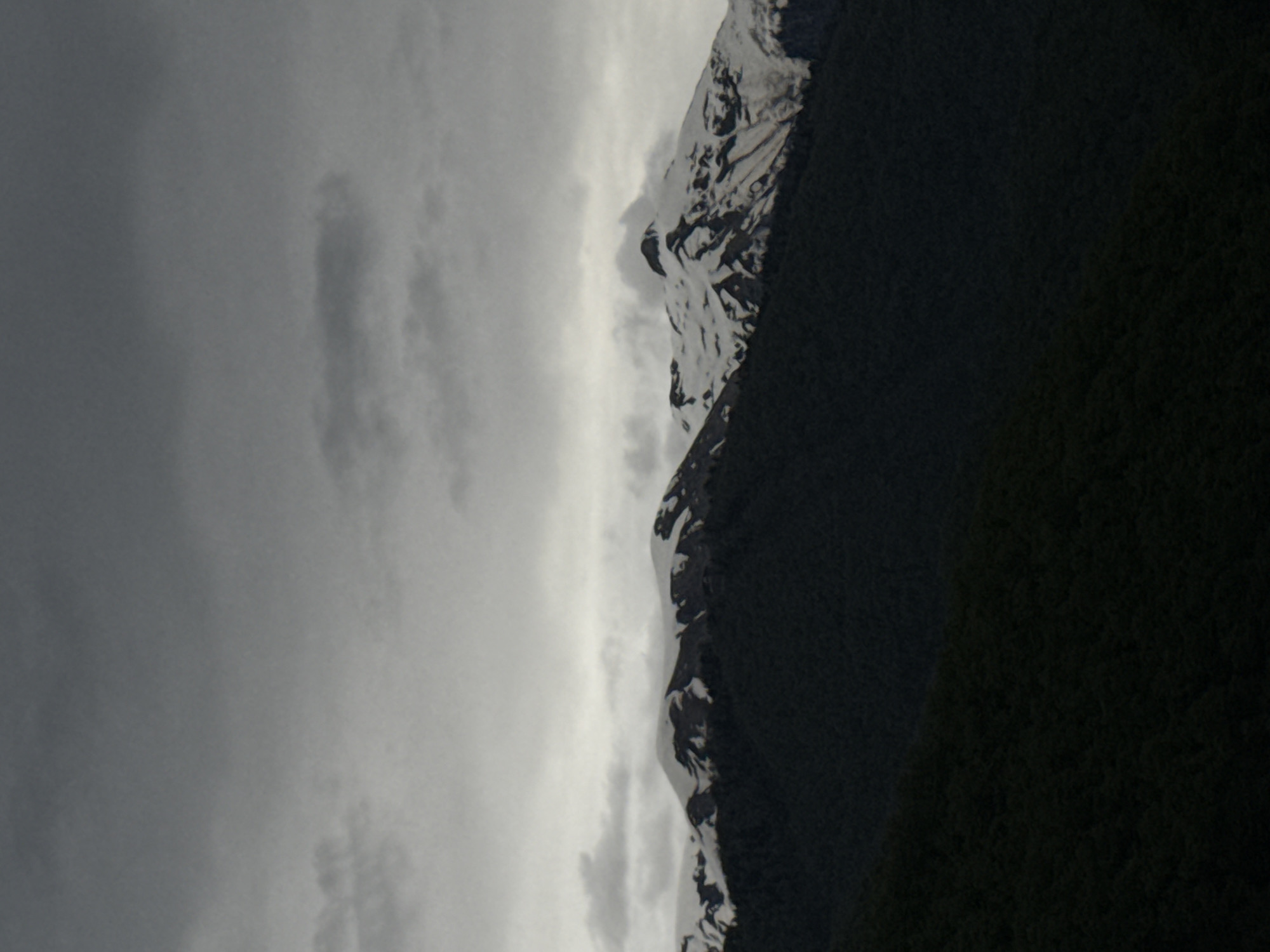 The image shows a mountain range under a cloudy sky. The mountain range has some snow on the peaks and is densely forested on its slopes. The left side of the image is filled with gray clouds. The mountain range occupies the right half of the frame. Given the details, it appears to be a view of mountains typically found in Krasnaya Polyana, Russia.