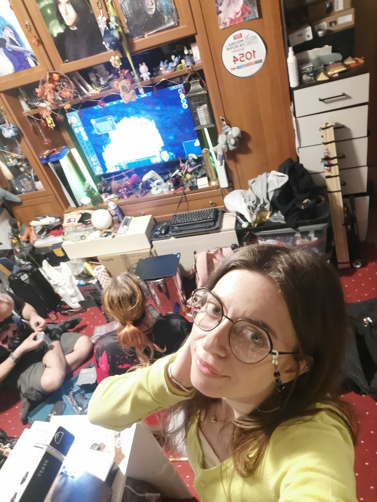 The image shows a person taking a selfie in a cluttered room. The room has a red patterned carpet and a wooden cabinet filled with various items, including figurines, posters of characters like Snape from Harry Potter, and a screen displaying a video game. A Playstation 5 sits vertically next to the screen. In the foreground, other people are sitting on the floor, seemingly engaged in some activity. Objects scattered around the room include boxes, electronics, and general clutter. A guitar stand is visible near a set of white drawers.
The presence of a "Я люблю Москву" sticker on the wooden cabinet indicates that the location is Moscow, Russia.