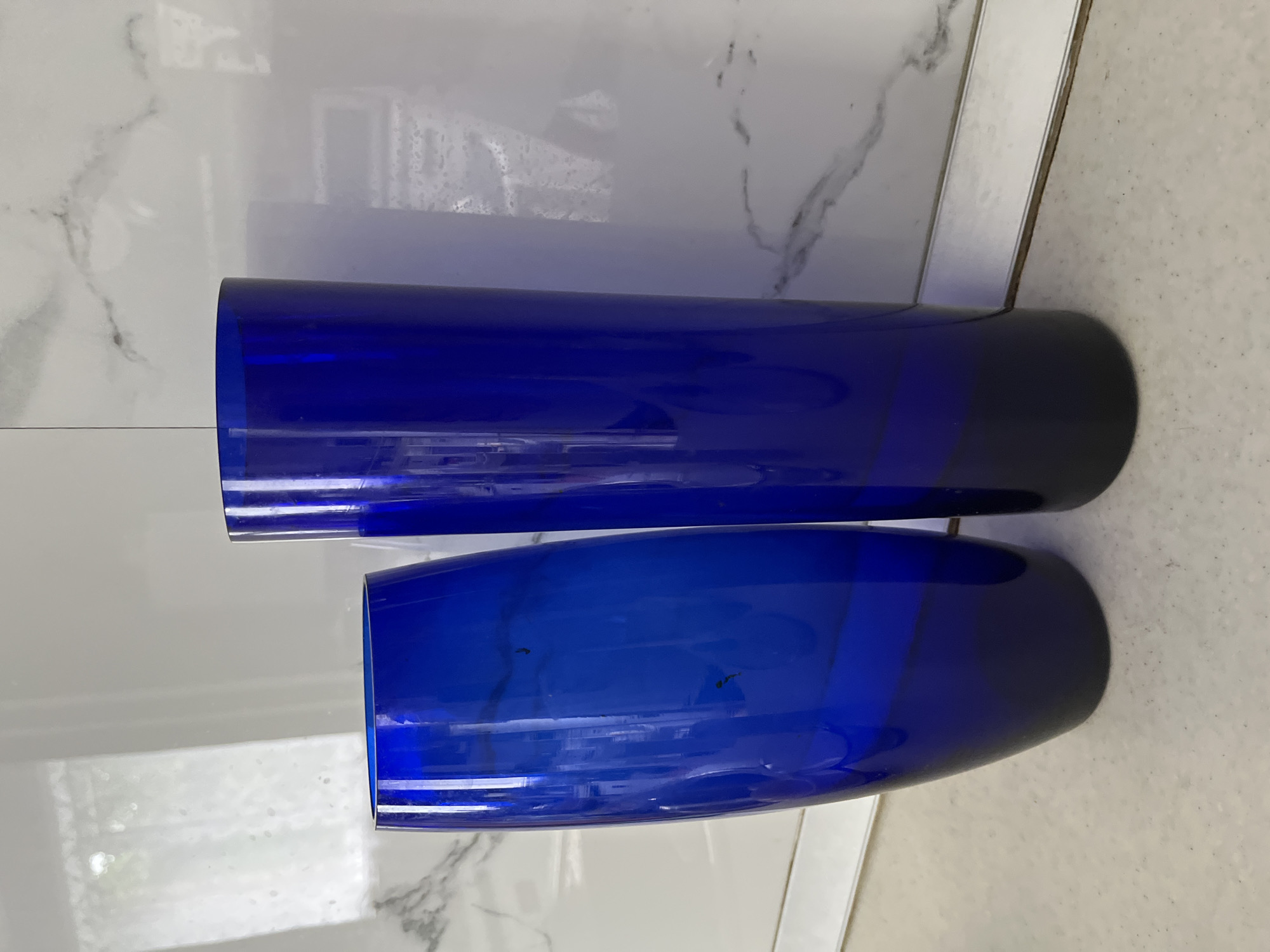 Two tall, cobalt blue glass vases are placed on a light-colored countertop. The vase on the left is wider and tapers towards the bottom, while the vase on the right is slender and cylindrical. Both vases have a dark, almost black, gradient at their bases. The background features a white tiled surface with subtle grey veining, resembling marble, and a reflective surface above that shows a blurred window and light. A thin silver strip runs horizontally along the bottom of the tiled area. There are no people or animals present. The scene is indoors, likely a domestic setting. The location is not specified.