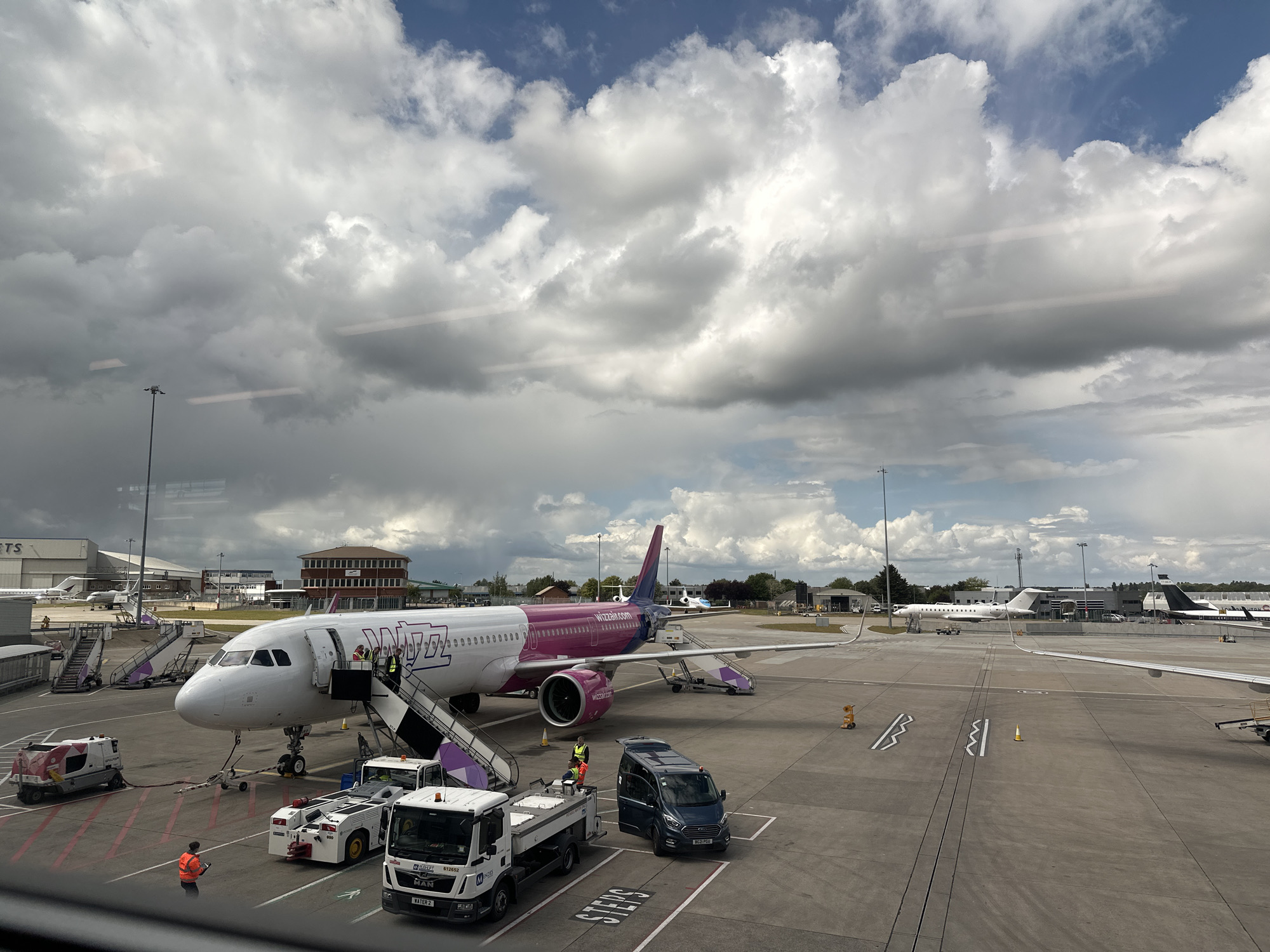The image depicts an airport scene with a Wizz Air Airbus A321 aircraft parked at a gate. The airplane is primarily white with a pink fuselage and tail. Passengers or crew members are seen near the boarding stairs. Various ground support vehicles are present, including a stair truck, a catering truck, and a van.
The location is an airport in the city of Unknown, Unknown. In the background, there are other aircraft, airport buildings, and the sky is filled with clouds. The photo is seemingly taken from an elevated position, likely inside the airport terminal.