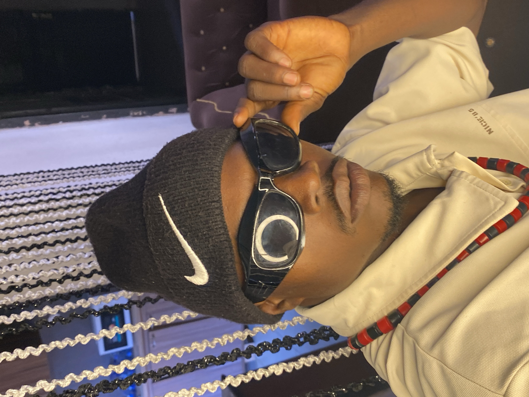 The image shows a close-up of a young Black man wearing a black Nike beanie, stylish black sunglasses, and a light-colored polo shirt. He is also wearing a red and black beaded necklace. The man is holding his sunglasses with his right hand. In the background, there are black and white beaded curtains and a glimpse of a dark room with purple furniture. The scene appears to be indoors, possibly in a home or studio. The specific location in Abakaliki, Nigeria, is not discernible from the visual information alone.