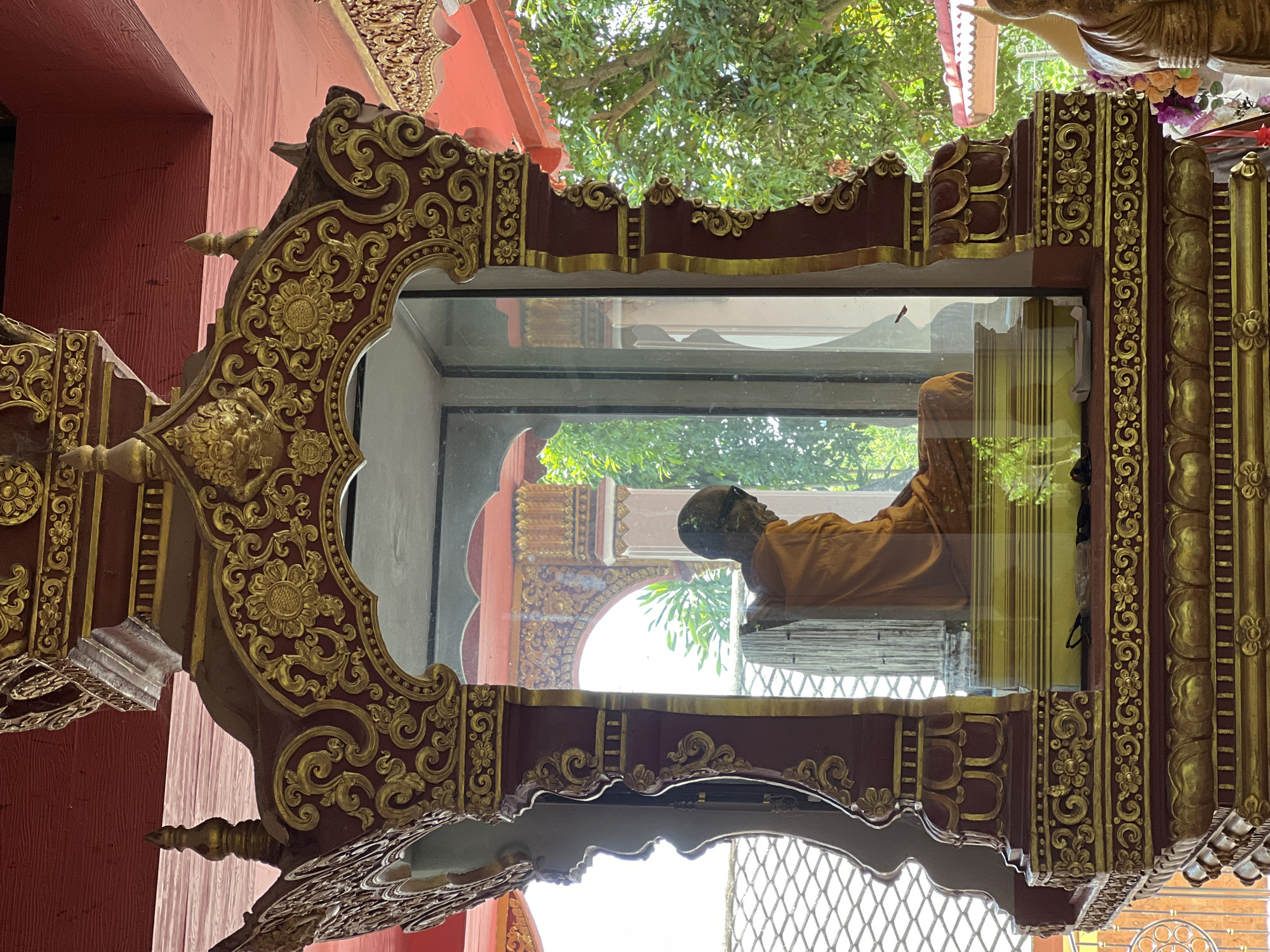 This is a full, eye-level, outdoor shot of a monk's mummified body encased in glass, inside a highly ornate, red and gold shrine at Wat Phra Non, a Buddhist temple located in Baan Hua Thanon, Thailand. The shrine is decorated with intricate gold carvings, featuring floral and swirling patterns. The monk, wearing an orange robe, is seated and appears to be in a meditative pose. The glass enclosure reflects the surrounding temple architecture, including an arched doorway with golden decorations, and lush green trees. The shrine itself is adorned with various decorative elements, including lotus petal motifs. The flooring within the enclosure is a metallic gold color. The roof of the shrine is a deep red, and the ceiling above is also red, with visible wooden beams. In the foreground, to the right, a golden Buddha statue is partially visible, adorned with a flower garland. The overall scene conveys a sense of reverence and cultural significance, typical of Buddhist temples in Thailand.