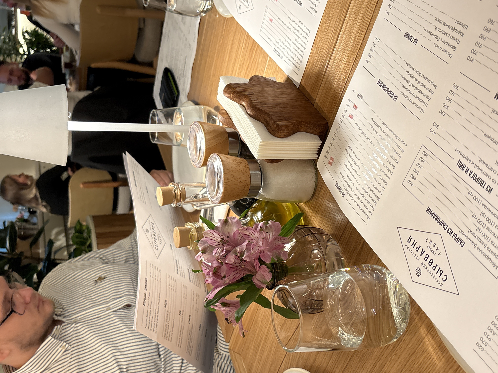 The image depicts a dimly lit interior of a restaurant, possibly in the city of Perm, Russia. In the foreground, a man wearing glasses and a striped shirt is seated at a wooden table, reading a menu. On the table, there are two glasses of water, a salt and pepper shaker set with wooden tops, a napkin holder filled with white napkins, a small vase with pink flowers, and two bottles of oil and vinegar. A portion of another menu is also visible. In the background, several people are seated at other tables, some of whom are blurred. The overall atmosphere appears to be relaxed and intimate.