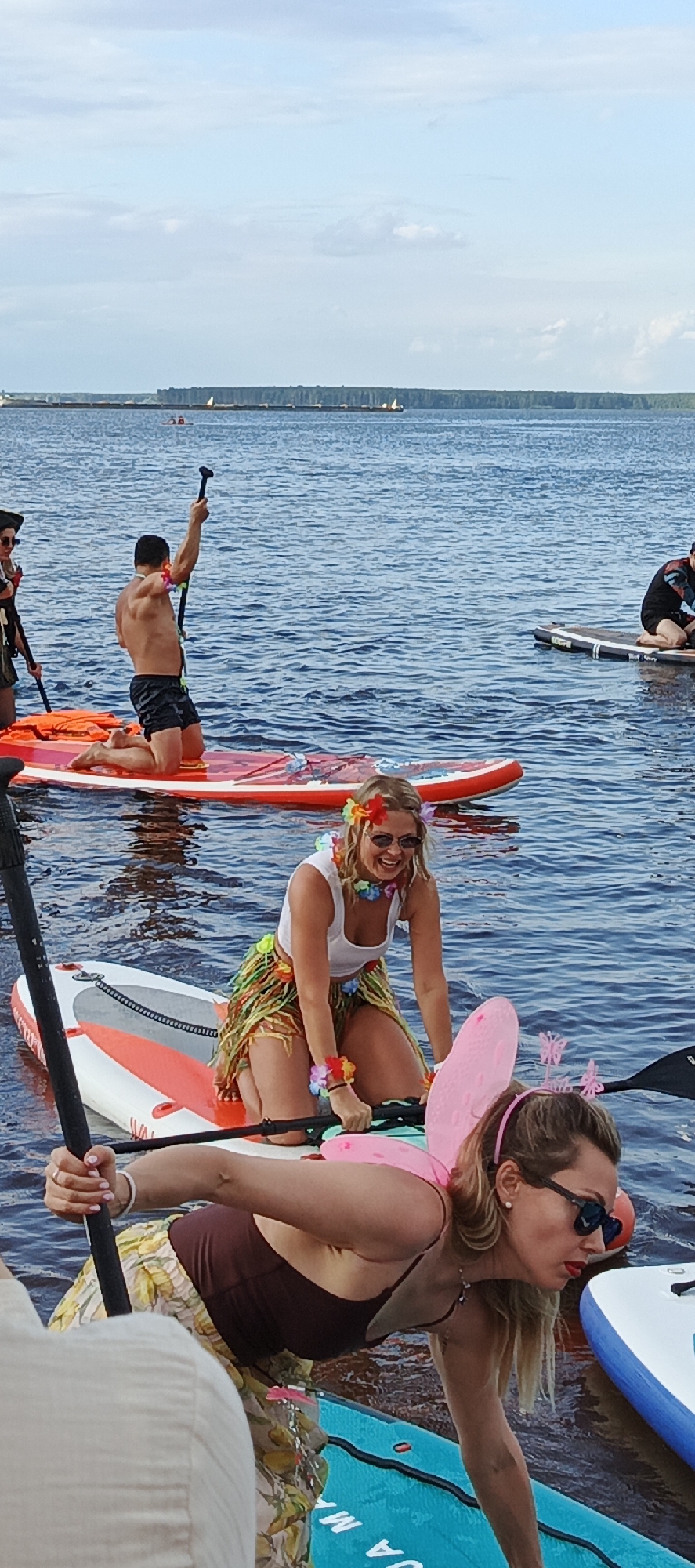 The image shows a group of people on paddleboards in a body of water, likely a river or lake. The scene appears to be a casual outdoor activity, possibly a themed event given the leis and a fairy costume with wings worn by one of the individuals. The water is calm and blue, with a hazy shoreline in the background and a cloudy sky above. Several paddleboards are visible, some with paddles. People are in various poses, with some kneeling and others leaning. The overall impression is a sunny day with recreational activity taking place on the water. Based on the provided crops that show a shoreline with trees and a distant man-made structure, this location could be the Kama River near Chaikovsky, Russia.