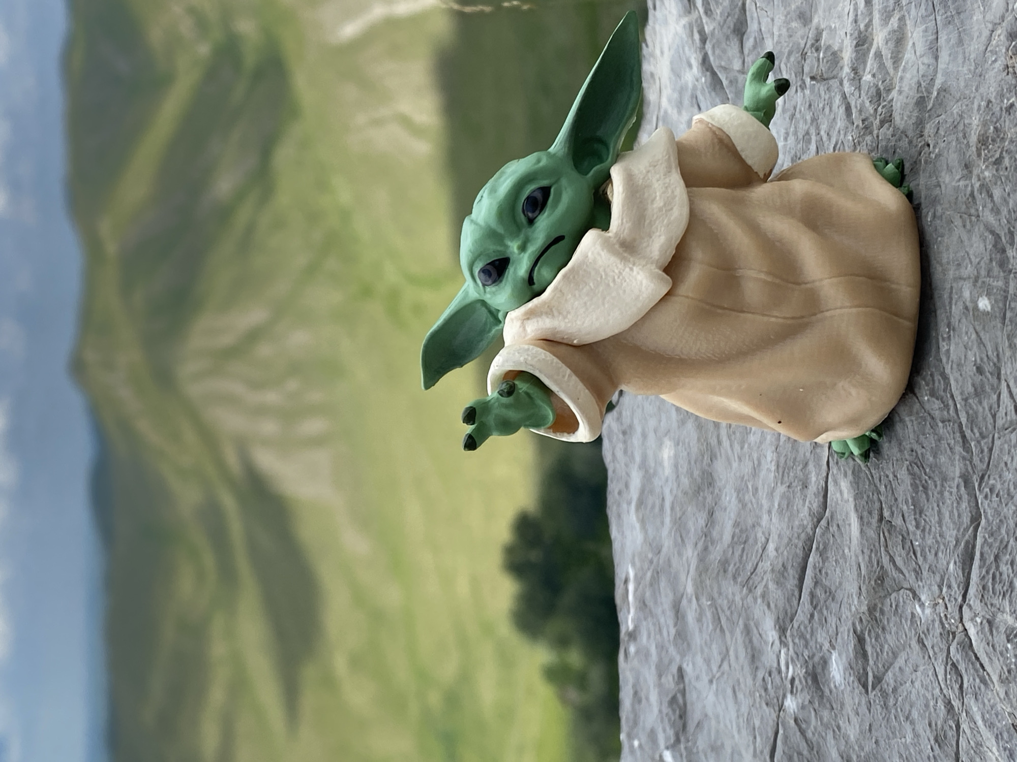 A figurine of the character Grogu, also known as Baby Yoda, stands on a rocky surface outdoors. The figurine is green with large ears and wears a tan robe with a cream-colored collar and trim. Grogu's left arm is extended forward, and its hand is visible. The background features a blurred landscape of rolling green hills under a cloudy sky. The specific location in Абзаново, Russia, cannot be determined from the image.