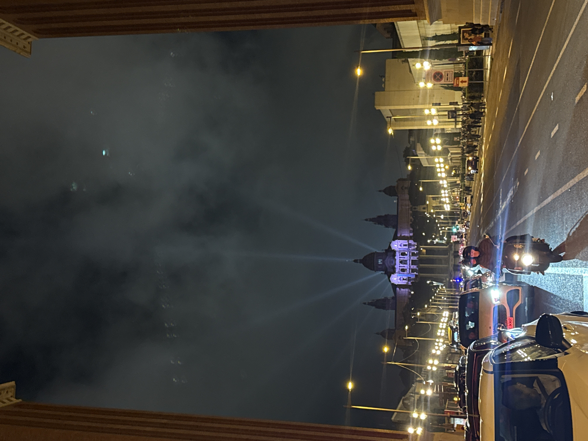 The image shows a night scene in Barcelona, Spain, featuring the Magic Fountain of Montjuïc and the Palau Nacional in the background. Light beams project upwards from the fountain. A street runs along the right side of the frame, with parked vehicles including cars and a motorcycle. Some people are visible on the street near the vehicles. The sky is dark, with visible atmospheric haze or mist.