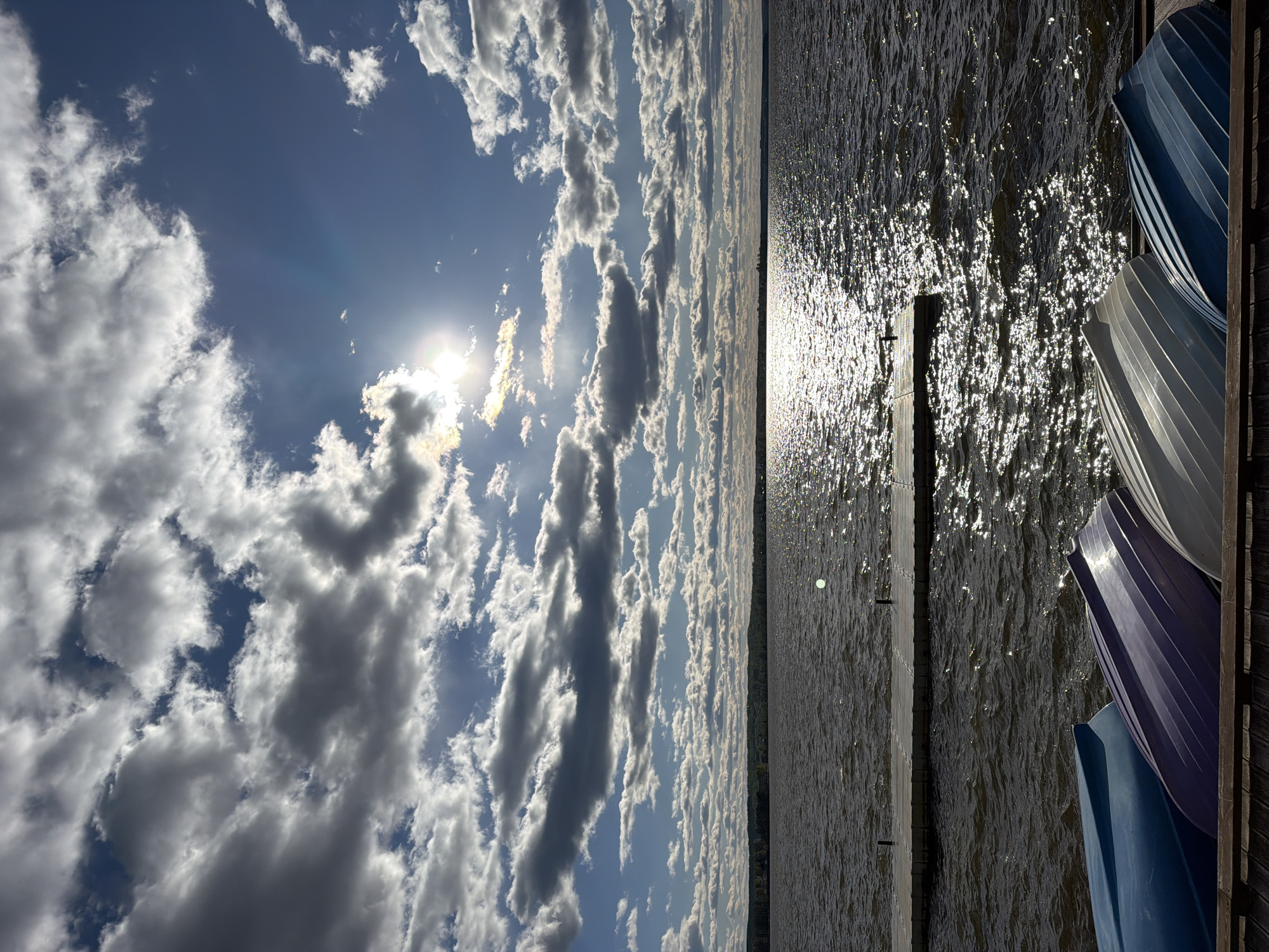 The image depicts a sunny day at a body of water, likely a lake or reservoir, possibly in or near Votkinsk, Russia. Bright sunlight glints off the water's surface, creating a sparkling effect. A wooden pier or dock extends into the water, and several small boats, likely rowboats, are stacked on a structure at the edge of the water. The sky is partially covered with cumulus clouds, with the sun visible through a break in the cloud cover. The angle of the shot seems to have been taken sideways. The scene is peaceful and reflects a natural environment.