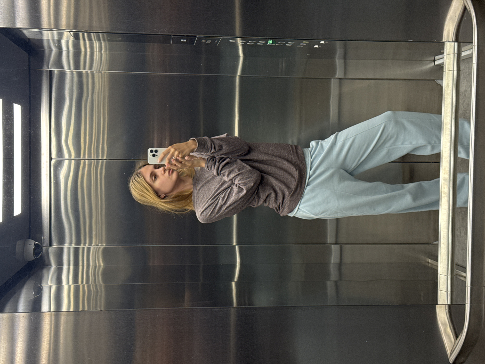 A woman with blonde hair is depicted standing in a modern elevator, taking a selfie using her white smartphone. She is wearing a dark gray long-sleeved top and light blue wide-leg pants. Both hands are holding the smartphone, which has three visible camera lenses on its back, as she angles it to capture her reflection in the highly polished, brushed metal walls of the elevator. Her expression is neutral, and her gaze is directed towards the phone's screen or lens.
The elevator interior features reflective stainless steel panels on all visible surfaces, showing distorted light streaks and reflections. Above the woman's head, an elevator control panel is visible, equipped with multiple buttons and indicators, including a small illuminated green light. A black security camera is mounted on the left wall of the elevator, near the top. On the right side of the image, a vertical metallic element, possibly a door frame or handrail, is visible. The scene is brightly lit, enhancing the reflective qualities of the metal surfaces. No distinct text is legible on the control panel or elsewhere within the image.