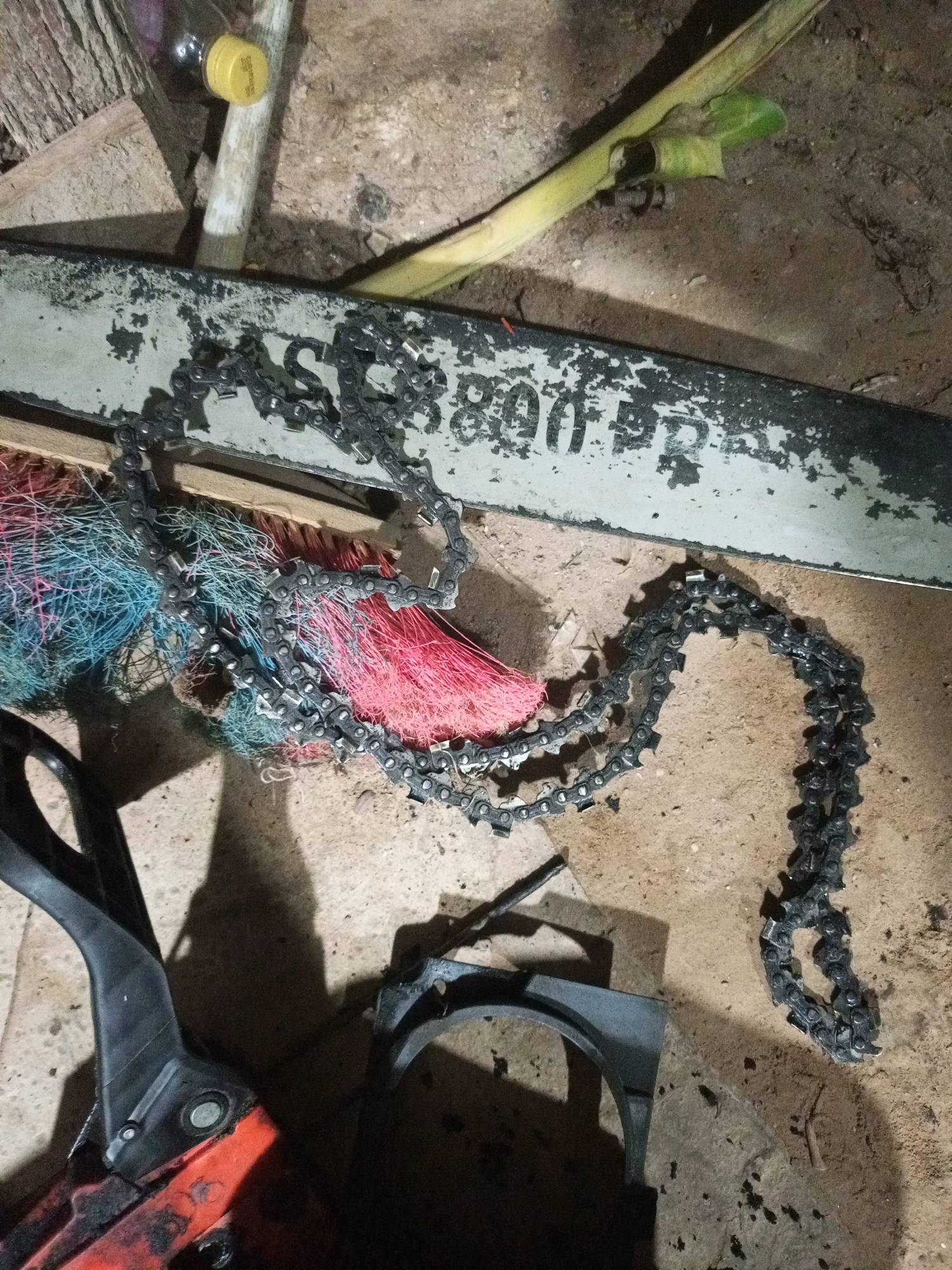 The image depicts a close-up, overhead view of chainsaw parts and debris scattered on a dirt or sand surface. A long, light-colored guide bar with faded black lettering, possibly reading "AS 3800 PR0," is positioned horizontally across the upper middle of the frame. A black chainsaw chain is draped over the guide bar and extends downwards. Beneath the guide bar, a bundle of multi-colored brush or bristles, predominantly pink and blue, is visible. In the lower left quadrant, the orange and black handle and part of the body of a chainsaw are visible, casting a shadow. A black plastic piece, possibly a chain guard, lies in the lower middle of the image. In the background, a yellow-capped bottle and what appears to be a piece of wood are present. The overall environment suggests an outdoor or workshop setting, with the presence of dirt and scattered materials. No people are visible in the image. The scene appears to be in Keling Panggau, Indonesia.