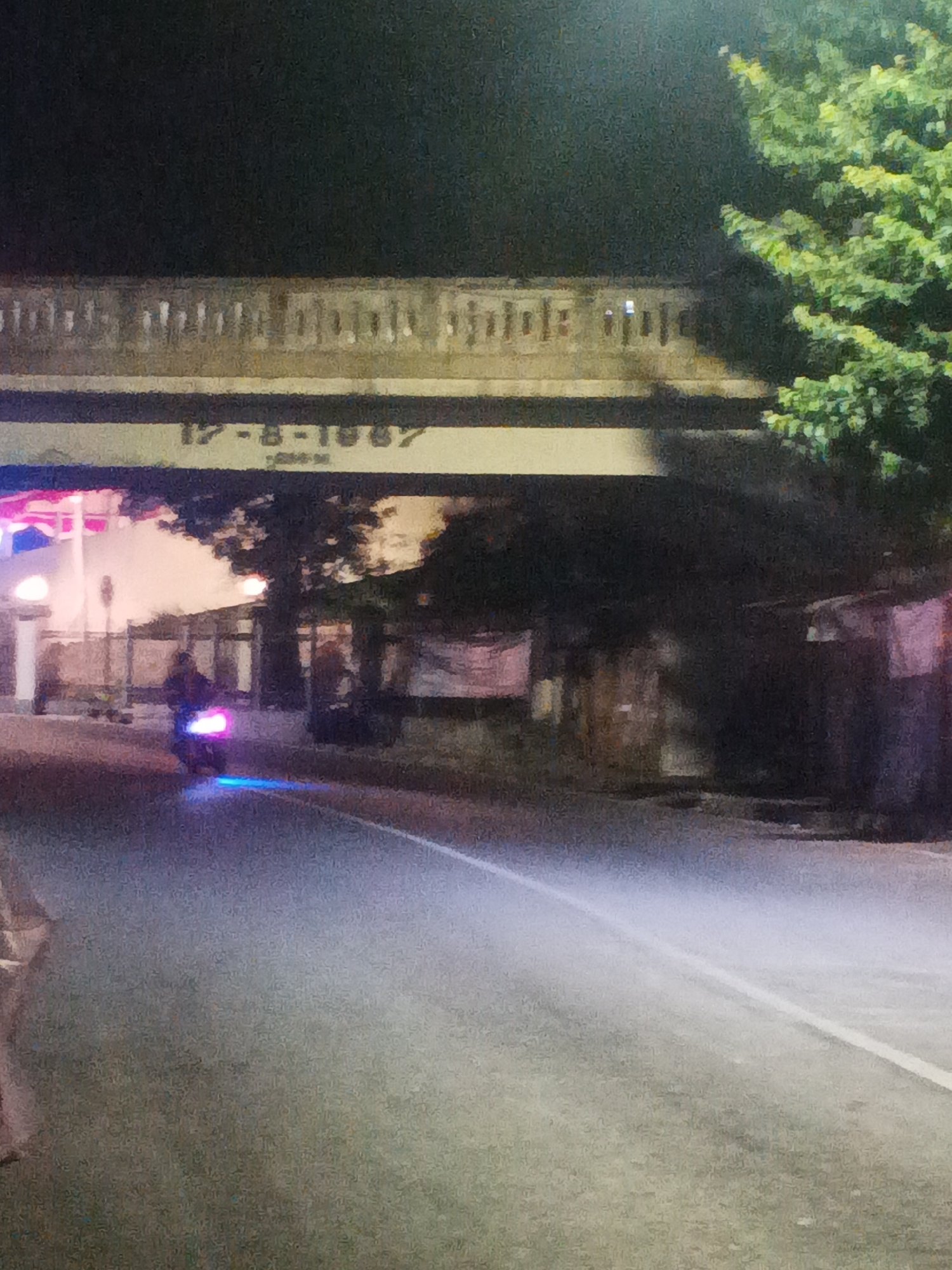 The image depicts an outdoor scene at night, likely in the city of Bantarsoka, Indonesia, as indicated by context not present in the image itself. A motorcycle, illuminated by bright pink and blue lights, is the central subject, moving on a paved road. The motorcycle's rider is silhouetted against the lights. Above the road, a bridge or overpass is visible with decorative elements and text that appears to read "17-8-1987". The background includes trees with green foliage and the soft glow of artificial lights, suggesting an urban or semi-urban environment. The overall atmosphere is dark, with the primary illumination coming from the motorcycle and the distant lights.