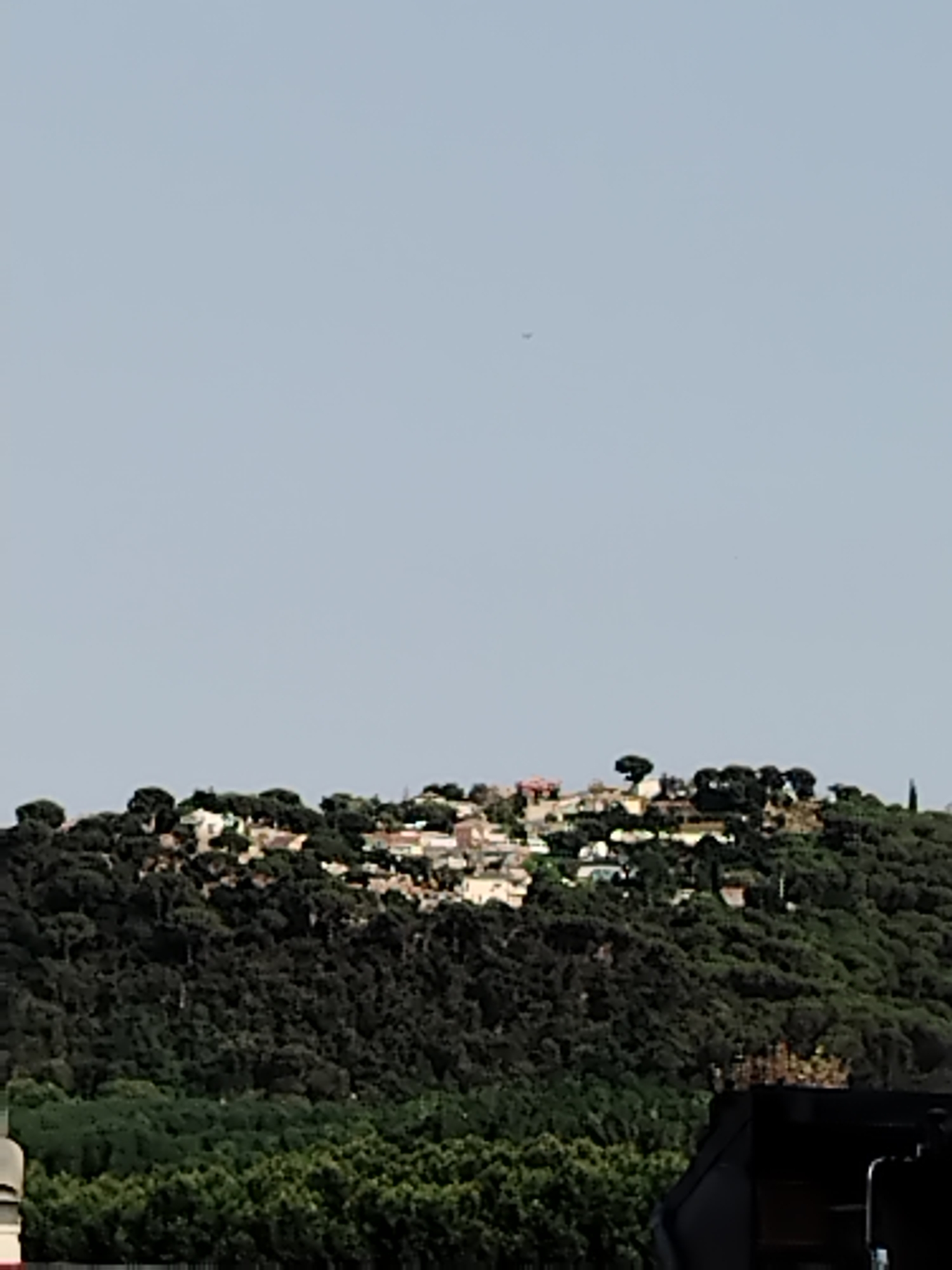 The image depicts a hillside covered in dense green trees, with a cluster of white buildings and red-tiled roofs situated on the upper slopes. The buildings appear to be houses or part of a village. The sky is a clear, light blue with a small, dark object, possibly an airplane, visible in the upper portion. The foreground shows more green foliage, suggesting a natural or park-like setting. There are no people or obvious actions taking place. The scene conveys a sense of a settled, elevated area within a natural landscape. This appears to be the viewpoint from a lower elevation looking up towards the town of Hostalric, Spain, which is known for its historical castle and situated on a hill overlooking the surrounding countryside.