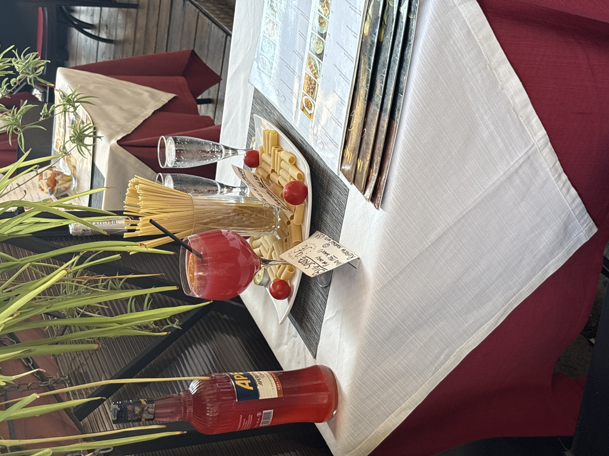 The image depicts an outdoor dining table at what appears to be a restaurant in Rome, Italy. The table is set with a white tablecloth over a maroon base. On the table, there is a bottle of Aperol, a glass of red cocktail with an orange slice, two glasses of sparkling water, and a plate of rigatoni pasta garnished with two cherry tomatoes. A small handwritten sign is placed next to the pasta, which reads "BIG SPOON: €5.00 for only 10.00 (and free snack!) REFRESS YOURSELF WITH US!". In the background, other tables with similar settings are visible, suggesting a dining establishment. The overall ambiance suggests a relaxed meal outdoors, possibly in the afternoon or early evening.