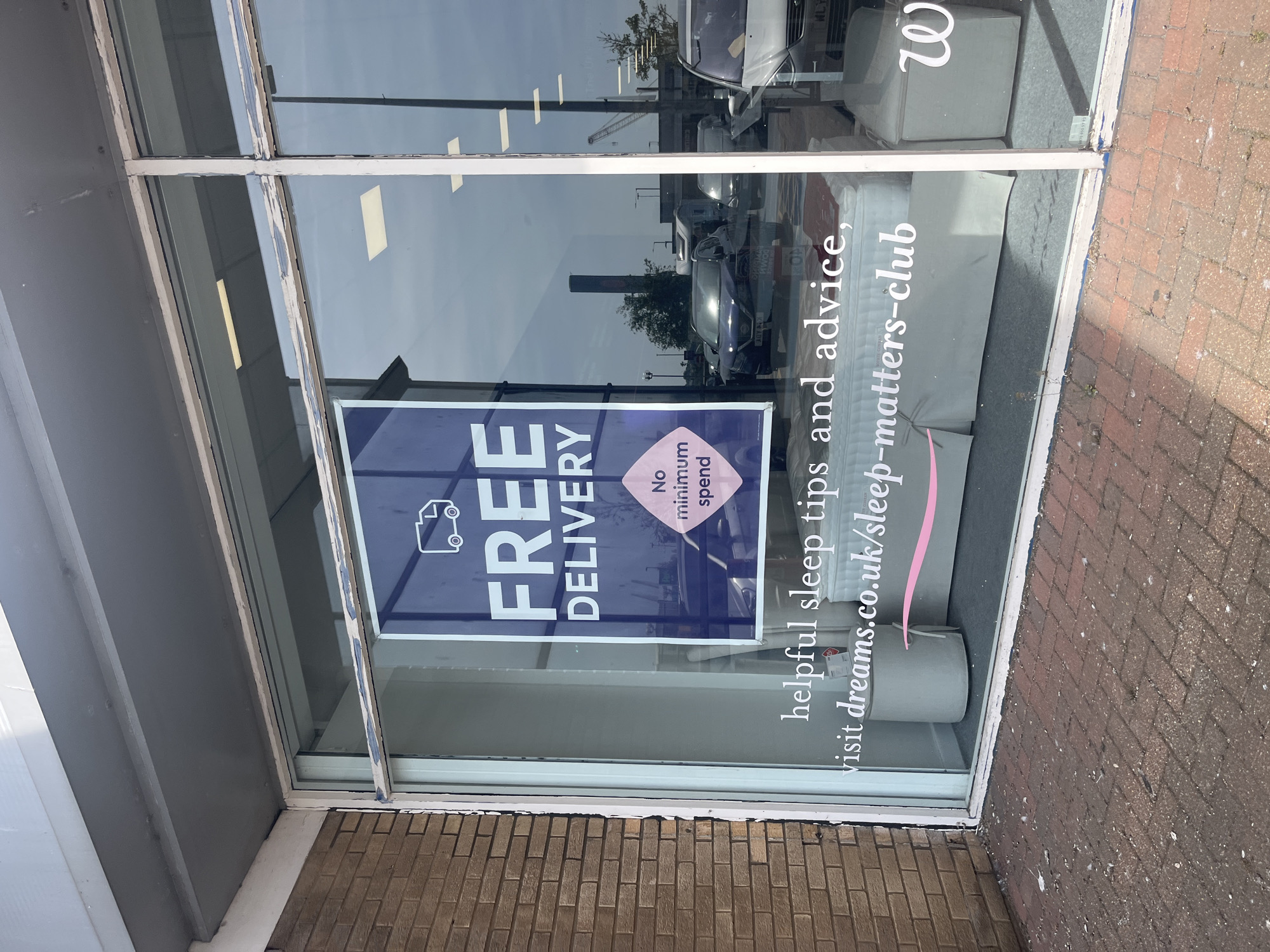 The image shows the storefront of a Dreams furniture store in East Suffolk, United Kingdom. A window display features a promotion for free delivery with no minimum spend. The store's website, dreams.co.uk/sleep-matters-club, is also advertised. The store is located on a street with cars and other commercial buildings. The facade of the storefront has a gray frame around the window, and there is a red brick wall visible near the bottom of the frame. The reflections in the window include street scenes and parked vehicles.