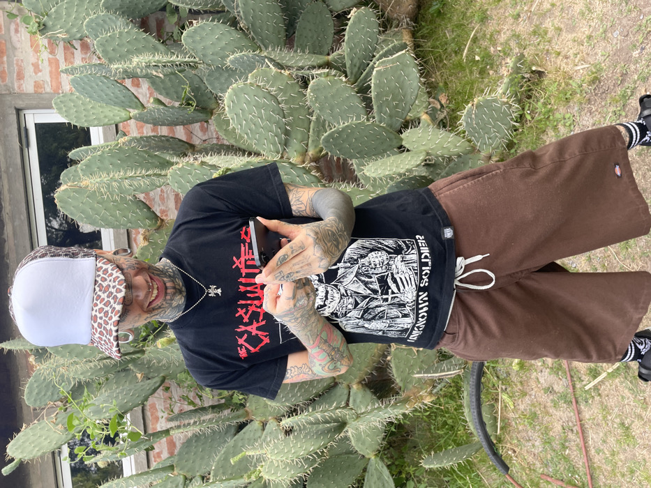 A lone individual stands outdoors in what appears to be a private garden or yard in a residential area of Buenos Aires, Argentina. The person, presenting as male, is heavily tattooed on the face, neck, arms, and hands, displaying prominent ear gauges, a septum piercing, and other facial piercings. They wear a white trucker hat with a leopard-print brim, a black short-sleeved t-shirt featuring the band name "THE CASUALTIES" in red and a white graphic of a skeleton labeled "UNKNOWN SOLDIER," brown cargo shorts with a white drawstring, striped socks, and black open-toed sandals. A silver chain with a cross pendant is visible around their neck. The individual holds a dark-colored smartphone with both hands, looking down at the screen, and exhibits a wide smile. The background consists of a dense cluster of green Opuntia cacti with visible spines, fronting a light-colored brick wall with a window. The ground is covered in grass, and a segment of a black tire or hose is partially visible in the lower-left foreground. The specific location within Buenos Aires is not discernible from the image.