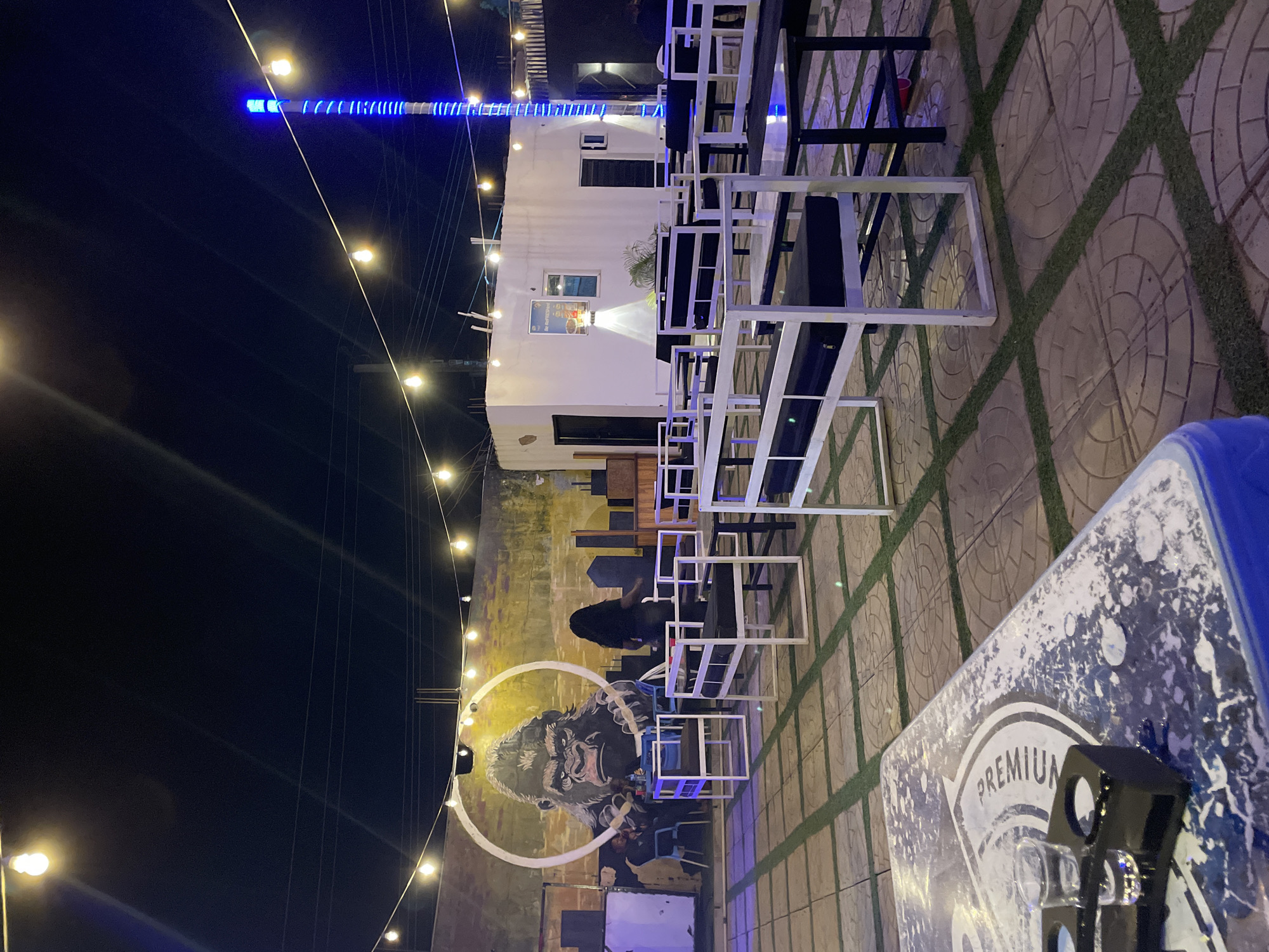 This is an outdoor scene at night, likely in a commercial area of Jos, Nigeria. Several rows of white metal chairs and black tables are arranged on a tiled surface with artificial turf in the seams. A mural of a gorilla's face is painted on a nearby wall. A string of lights is visible along the edge of the mural. A white, container-like structure with a window is in the background. A blue light is visible at the top of the structure. A table with a "PREMIUM" logo is in the foreground, and a drink dispenser is on the table. An individual is sitting at one of the tables with white chairs. Several other people are near the wall and the mural.