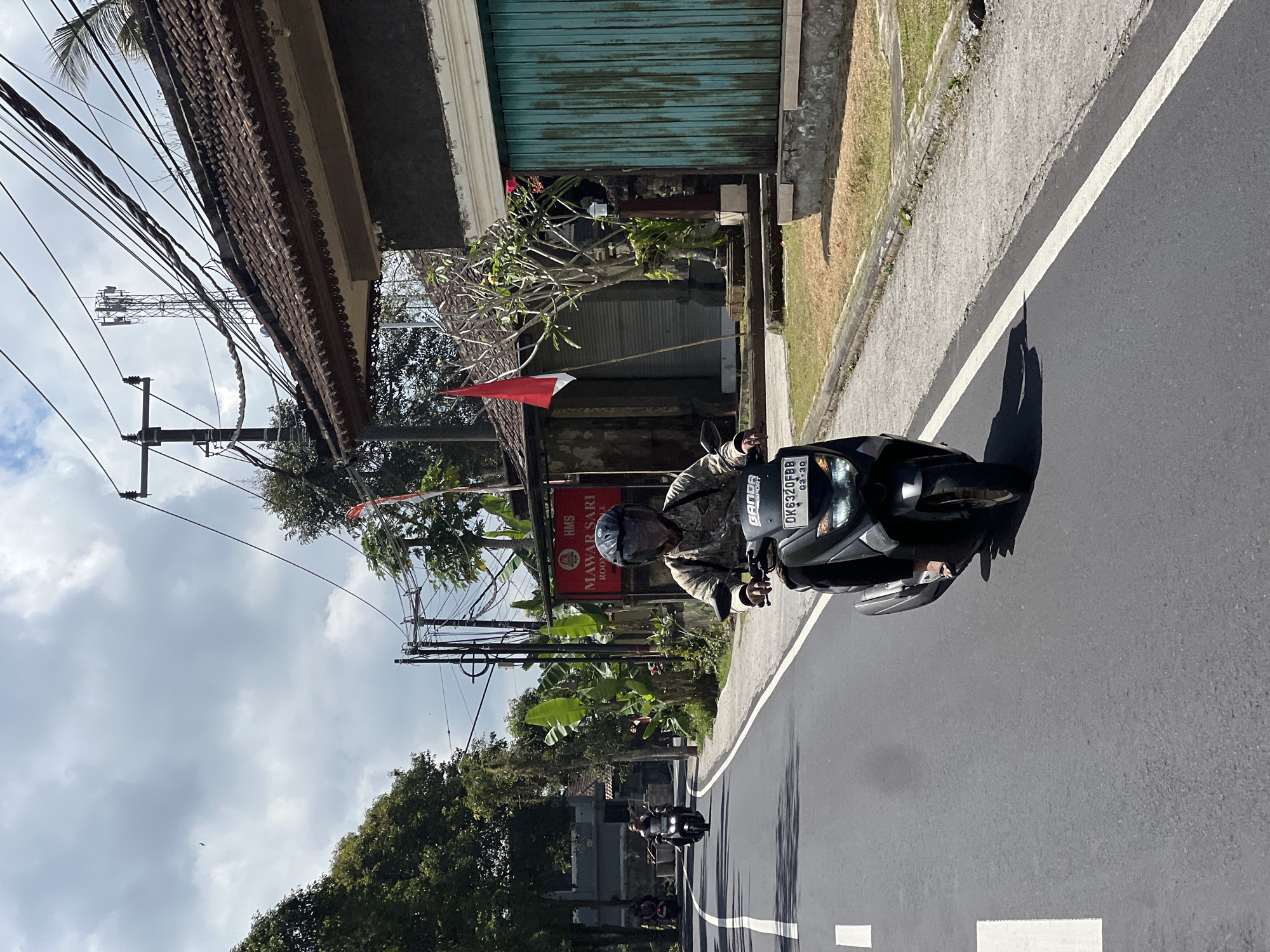 In Payangan, Indonesia, a person on a scooter rides down a paved road. The rider, wearing a camouflage shirt and a black helmet, is in the foreground. The scooter has a license plate that reads "DK6320FBB". In the background, there is a building with a red and white flag and a turquoise metal door. Electrical wires and a cell tower are visible in the sky. Other scooters are also seen on the road. The weather appears to be sunny with some clouds.