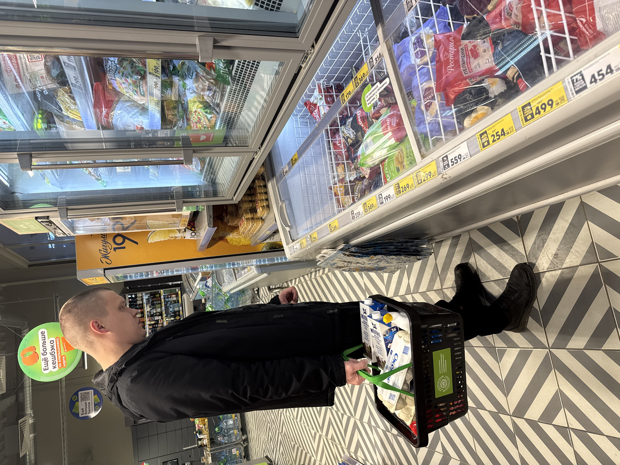 The image shows a man in a store, likely in Dmitrov, Russia. He is standing in an aisle in front of open freezer display cases containing various frozen foods. The man is wearing a black jacket and black pants, and black boots. He is holding a black shopping basket filled with groceries, including milk. The prices of the frozen food items are displayed on the shelves. The floor is tiled with a geometric pattern of grey and white tiles. Overhead are signs advertising a loyalty program with a lime green discount tag, reading "Eщё больше кешбэка".