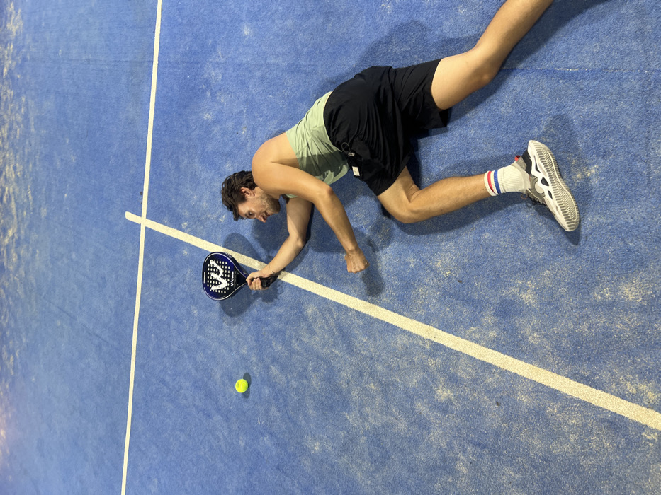The image displays a male subject on a blue padel court in Pecatu, Indonesia, under artificial illumination. The court surface, visible from an overhead perspective, exhibits wear and is marked with white lines. The subject is positioned on the ground, extending from the lower right to the upper left of the frame. He wears a light green sleeveless tank top, black athletic shorts, white athletic socks with red and blue stripes, and grey and white athletic shoes. His right arm is outstretched, holding a dark padel racket with a white 'VW' or 'VV' logo on its face. A yellow-green padel ball is located to the left of the racket. The subject's head is turned towards the ball, and his facial expression suggests exertion. His left hand is clenched into a fist at chest level, indicative of a dynamic athletic maneuver.