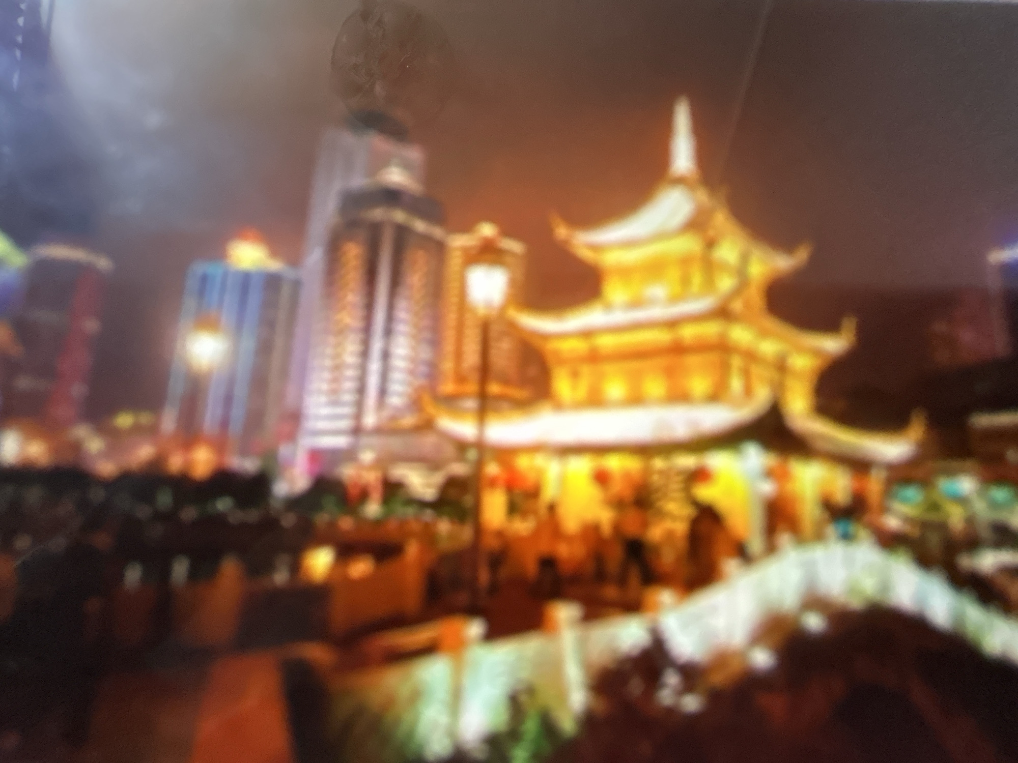 The image depicts a nighttime scene in Gelendzhik, Russia, featuring a traditional East Asian-style building illuminated with golden lights. This architectural structure, with its tiered roofs and ornate details, stands prominently in the foreground. To its left, modern skyscrapers with illuminated windows rise into the dark sky. A streetlamp with a classic design is visible between the traditional building and the skyscrapers, casting a warm glow. In front of the illuminated building, a white bridge with railings spans a dark area, possibly a body of water or a garden. The scene is busy with blurred figures of people walking and gathering, suggesting a public space or an event. The overall atmosphere is lively and festive, enhanced by the bright lights and the sense of activity. The time of day is clearly night, indicated by the darkness of the sky and the prominence of artificial lighting. There is no visible text in the image.
